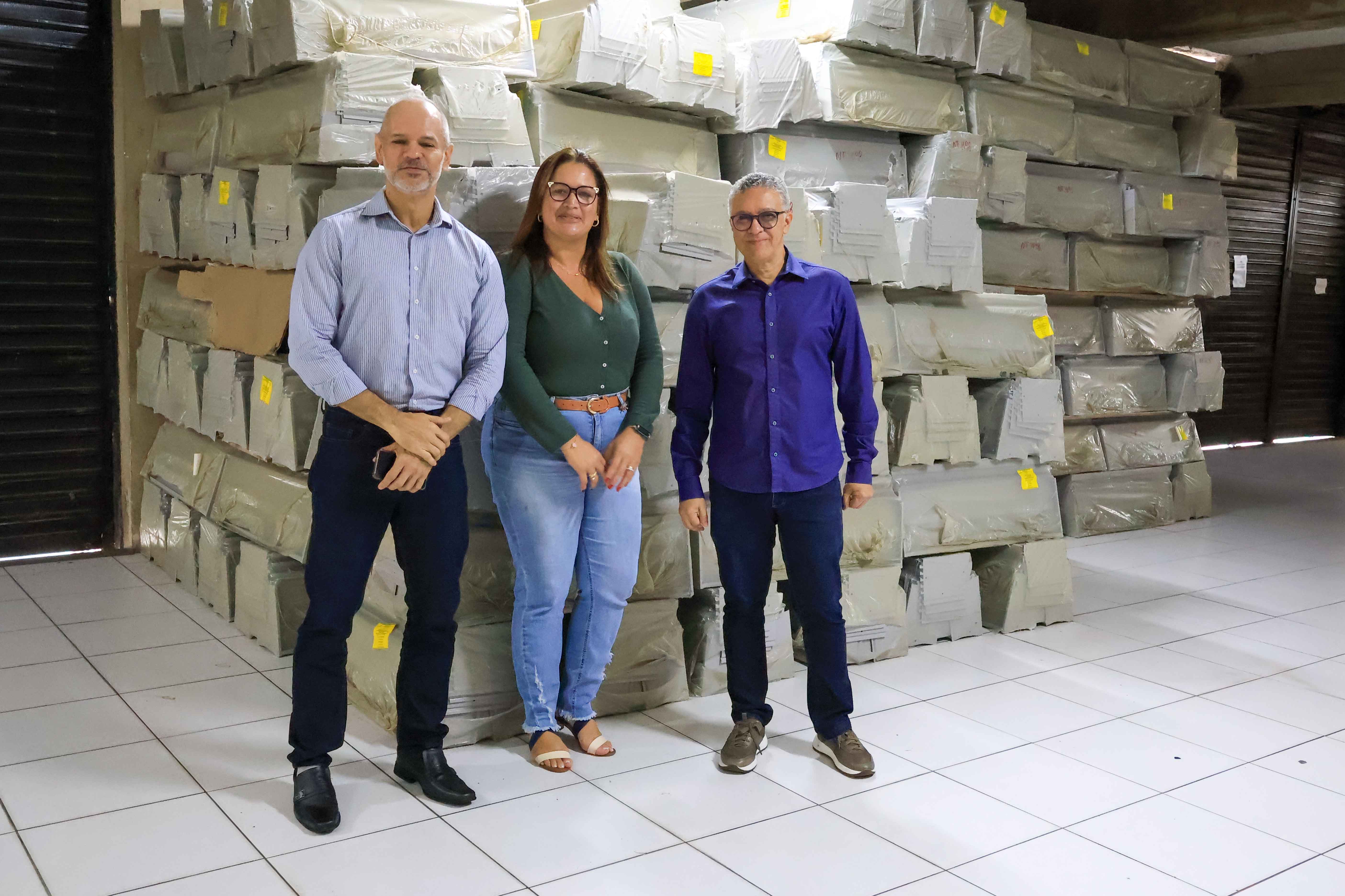 A imagem mostra três pessoas em frente a uma grande pilha de materiais embalados, organizados em um ambiente fechado. Os pacotes estão identificados com etiquetas amarelas, indicando um possível processo de catalogação ou distribuição. 