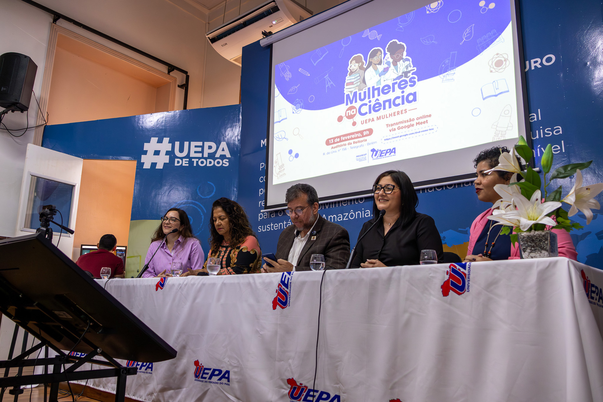 A foto mostra uma mesa de autoridades durante um evento institucional realizado em auditório da Universidade do Estado do Pará (UEPA). Cinco pessoas estão sentadas lado a lado atrás de uma mesa coberta com toalha branca com o logotipo da universidade, cada uma com microfone e copo de água à frente. Ao fundo, um telão projeta a identidade visual do evento “Mulheres na Ciência – UEPA Mulheres”, com ilustrações e informações de data e transmissão online.  O cenário inclui um painel azul 