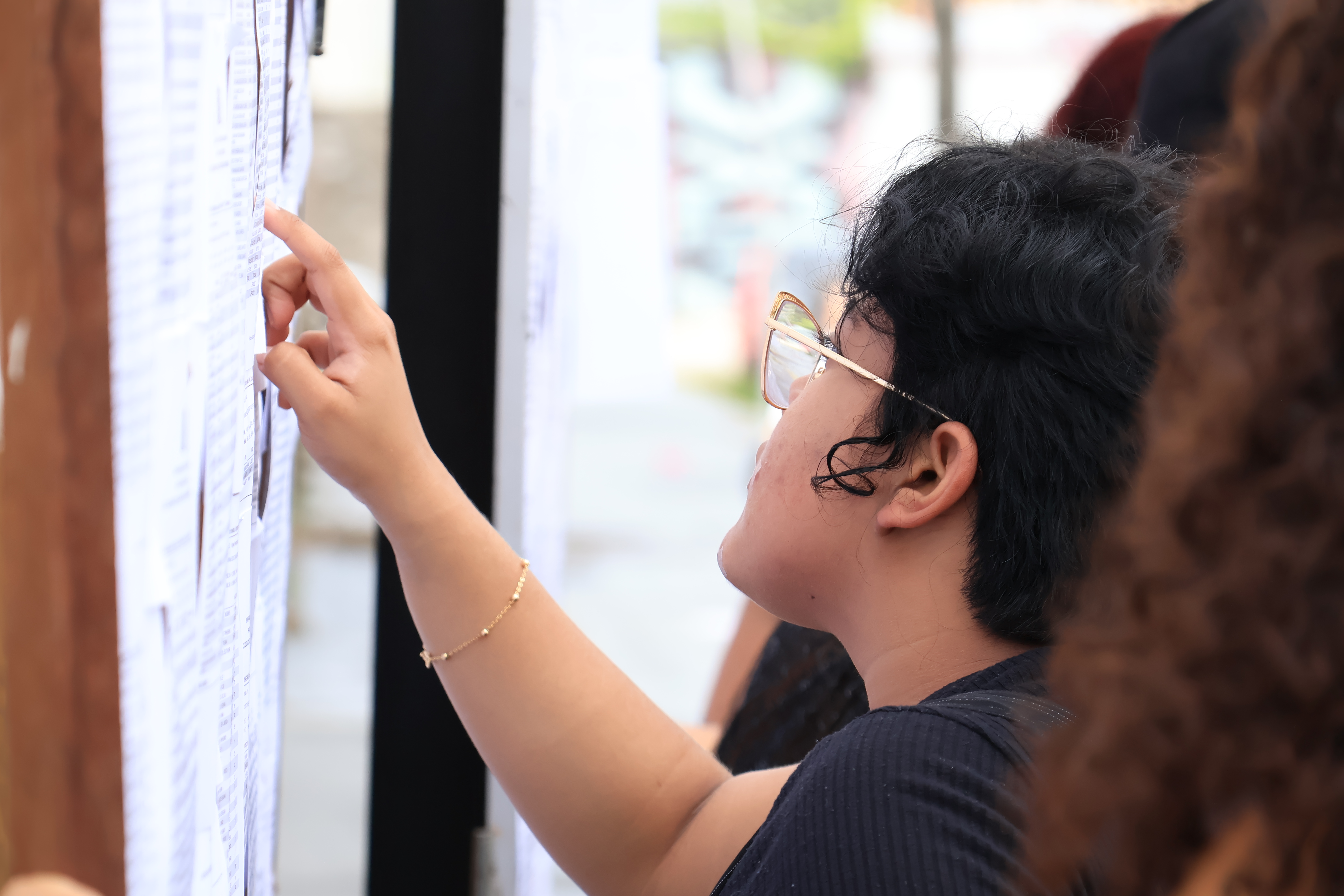 A imagem mostra uma pessoa de cabelos curtos e cacheados, usando óculos e uma blusa preta, apontando para um painel repleto de folhas com textos impressos. Ela parece estar procurando alguma informação específica na lista. O ambiente sugere um momento de divulgação de resultados ou informações importantes, possivelmente de um vestibular ou concurso público. Outras pessoas estão ao redor, também observando o painel. O foco da imagem está na pessoa em primeiro plano, com um fundo desfocado.