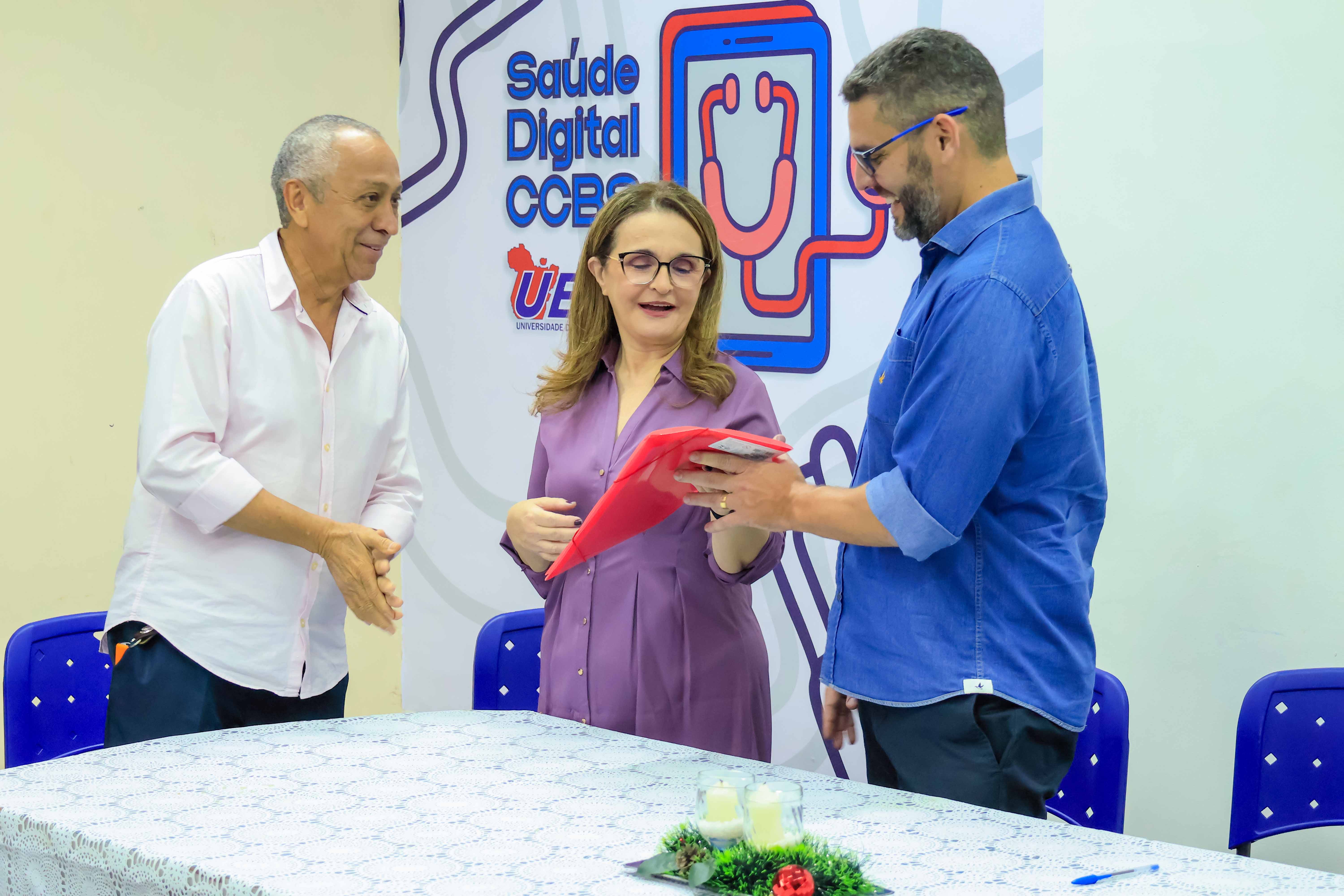 A foto mostra três pessoas em um evento da Universidade do Estado do Pará, como indicado no banner ao fundo que menciona "Saúde Digital CCBS". Há uma mulher no centro, vestida com uma blusa roxa, segurando um objeto vermelho enquanto conversa com um homem à direita, que usa óculos e uma camisa azul. À esquerda, um homem mais velho, vestido com uma camisa branca, observa e sorri. Uma mesa coberta com toalha branca está à frente deles, decorada com um arranjo natalino e duas velas.