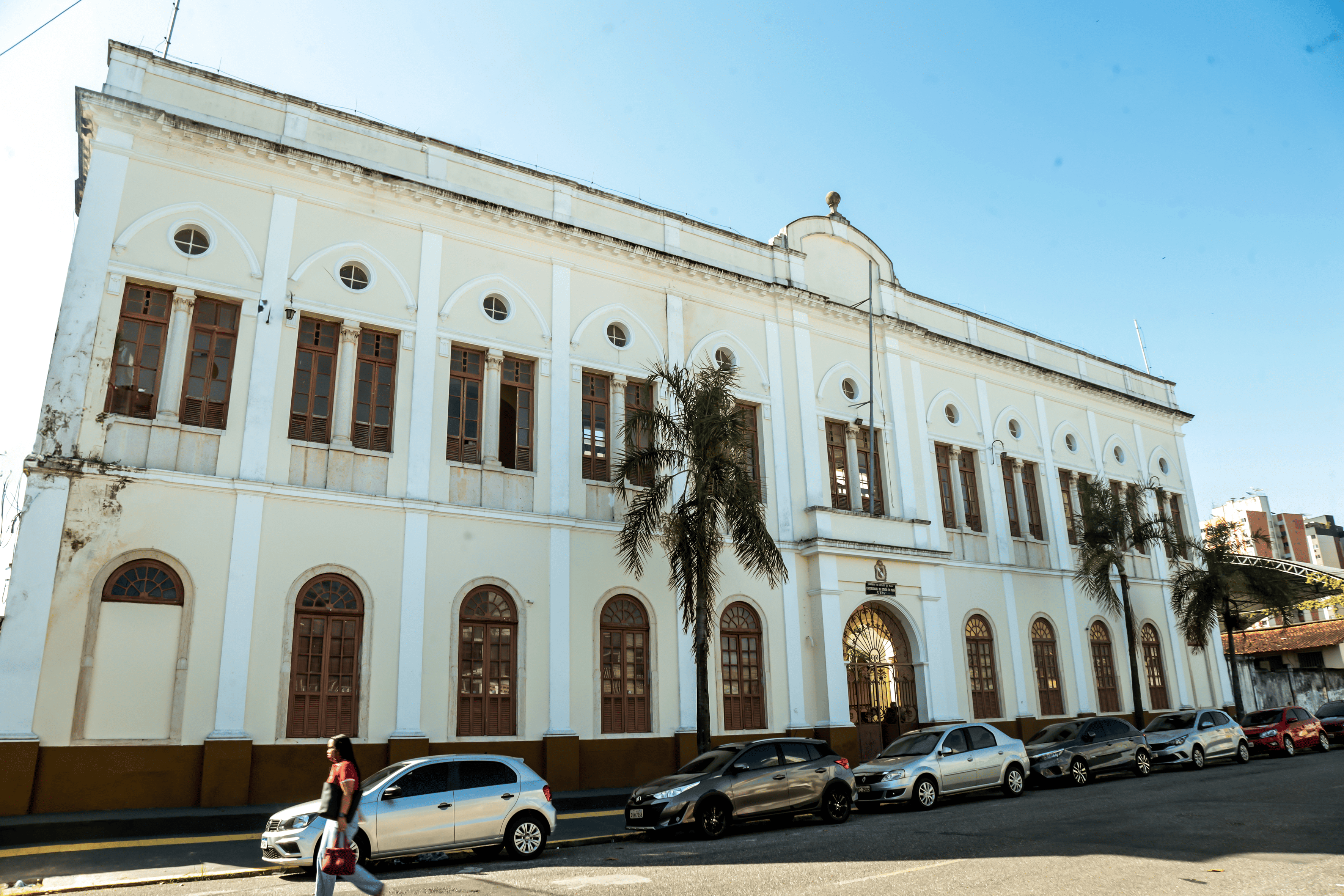 A imagem mostra a fachada de um prédio histórico de dois andares, com arquitetura clássica, pintado em tons claros e com detalhes em branco. O edifício possui janelas altas com molduras em arco e uma entrada central com portão ornamentado. Em frente, há algumas palmeiras e carros estacionados ao longo da rua, além de uma pessoa caminhando pela calçada em um dia ensolarado, com céu azul ao fundo.