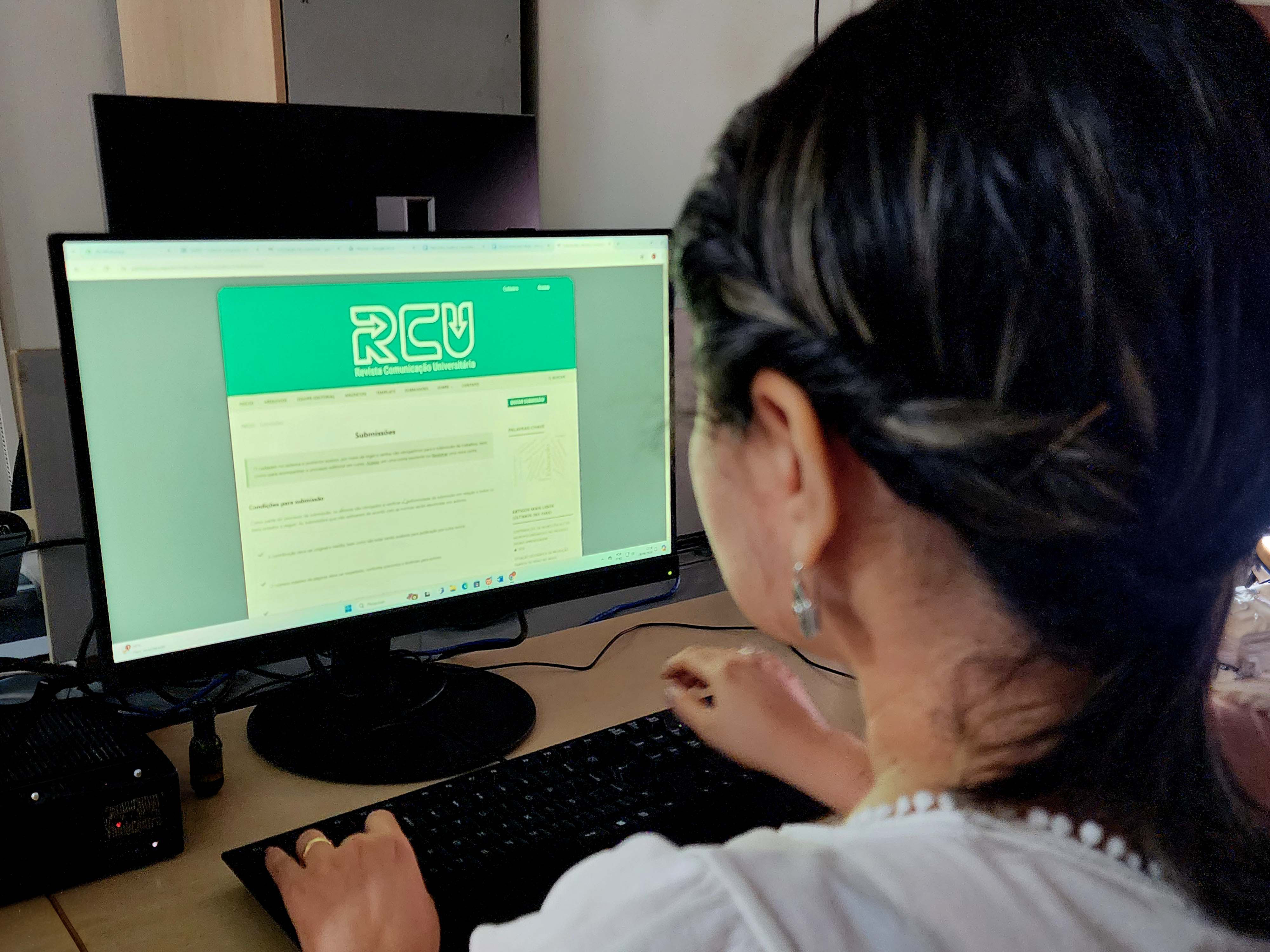 A imagem mostra uma pessoa vista de trás, olhando para uma tela de computador que exibe uma página da web com o logotipo “RCU” em letras grandes na parte superior. O conteúdo da tela mostra a home da Revista e retrata uma mulher interagindo com uma plataforma ou serviço online, para fins educacionais ou profissionais.