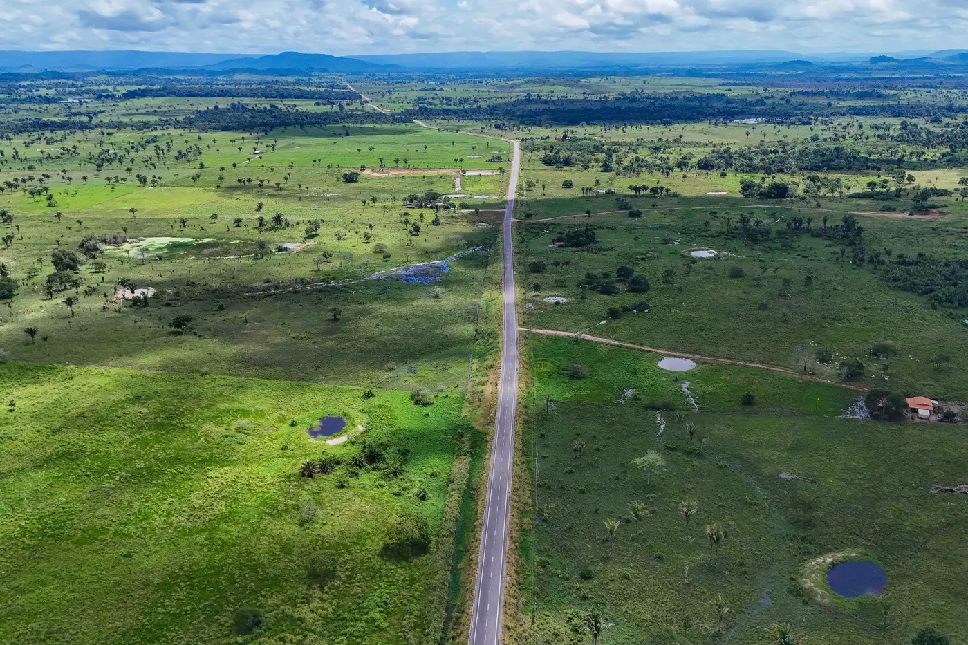 A imagem mostra uma paisagem rural ampla com uma estrada asfaltada cortando o campo em linha reta. O terreno é predominantemente verde, com áreas de vegetação esparsa, pequenos corpos d’água e algumas construções isoladas. Ao fundo, é possível ver elevações e serras sob um céu parcialmente nublado. A vista aérea destaca a imensidão da paisagem natural.