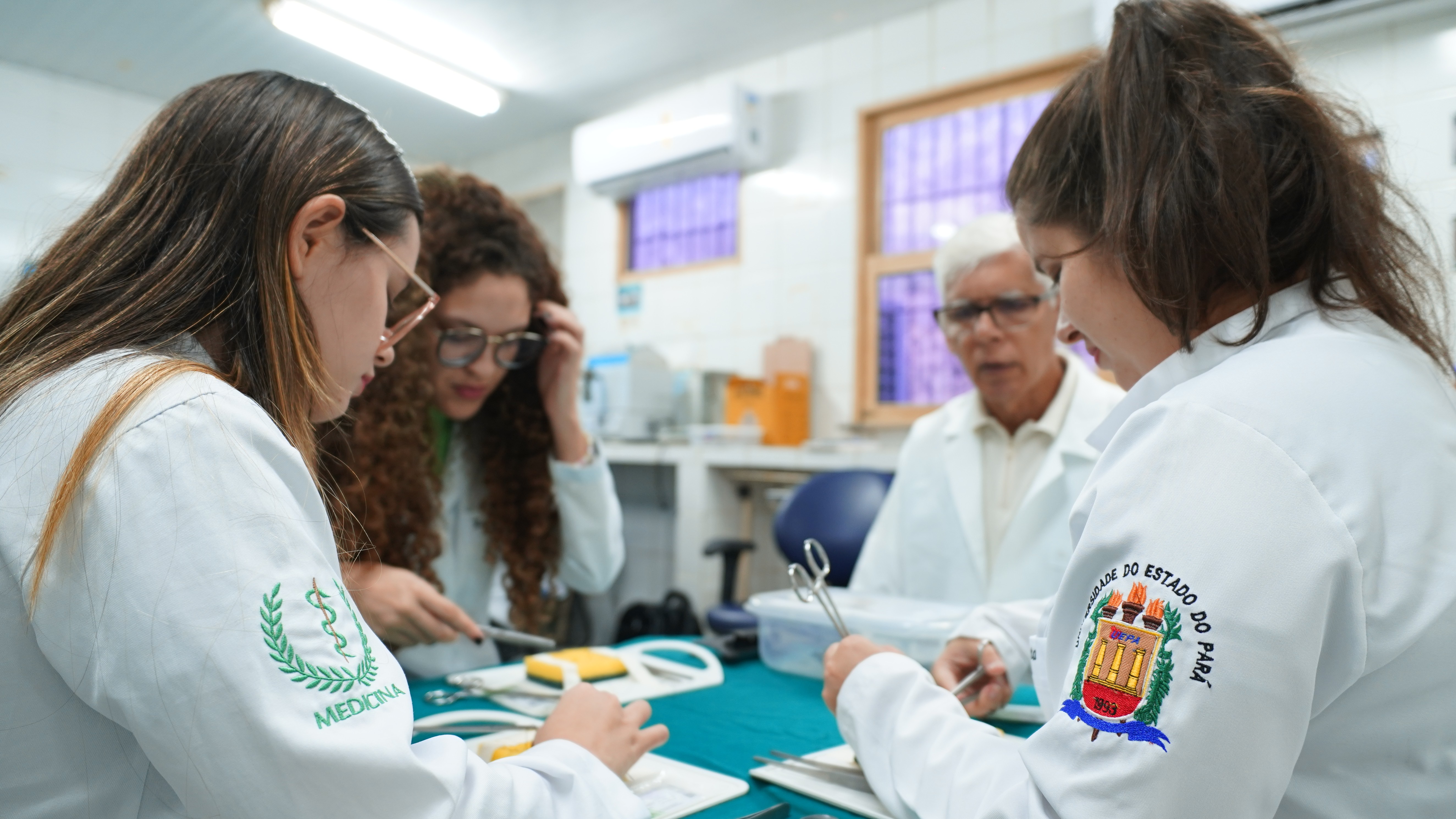 A imagem mostra um grupo de estudantes de Medicina da Universidade do Estado do Pará (Uepa) em atividade prática em laboratório. Eles estão usando jalecos brancos e manuseando instrumentos cirúrgicos sob a orientação de um professor, em um ambiente que simula uma sala de cirurgia experimental.