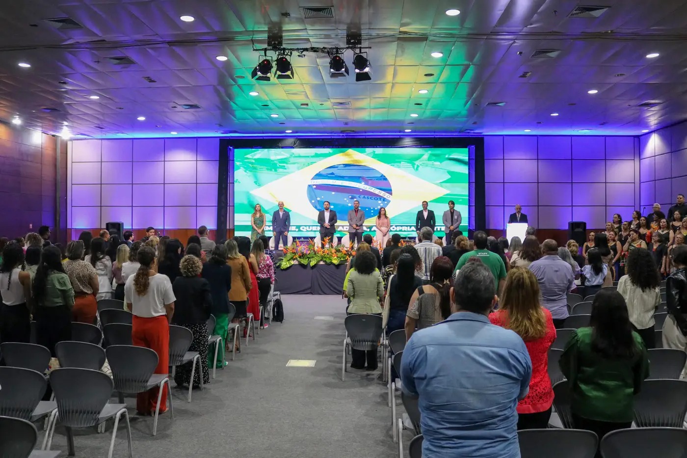 A imagem mostra um auditório lotado durante uma cerimônia formal. No palco, oito pessoas estão em pé diante de uma grande tela com a bandeira do Brasil. O público, também em pé, acompanha o momento solene. O ambiente é iluminado com luzes coloridas e decorado com arranjos florais à frente do palco.