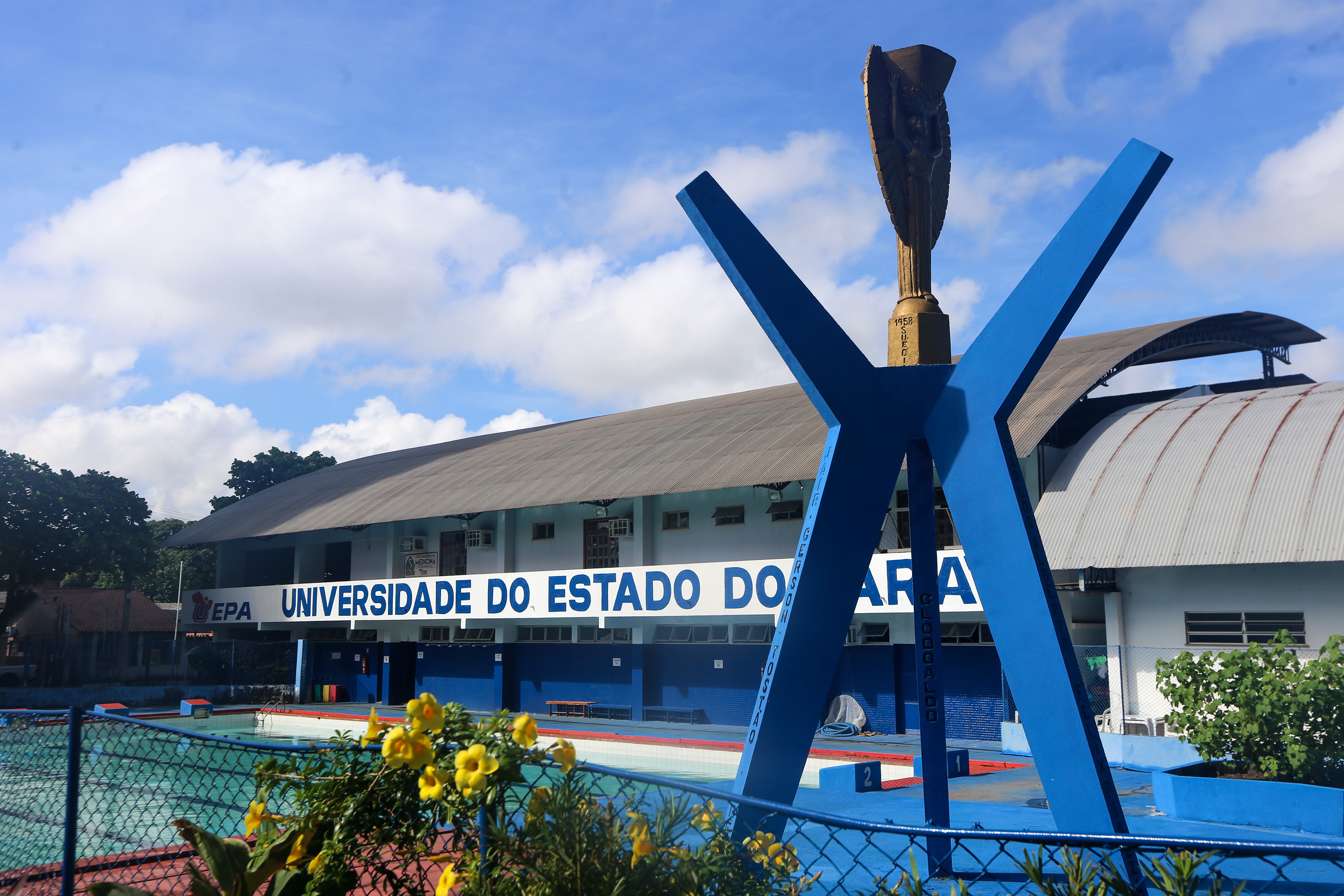 A imagem mostra a fachada de uma unidade da Universidade do Estado do Pará (Uepa). Em destaque, está o letreiro "UNIVERSIDADE DO ESTADO DO PARÁ" em letras grandes e azuis sobre a estrutura do prédio. No primeiro plano, há um monumento com uma escultura dourada no topo, sustentada por uma base em formato de "Y" pintada de azul. Ao redor do monumento, há uma área com piscina e gradil, com algumas flores amarelas visíveis no canto inferior esquerdo.  A arquitetura do prédio apresenta um telhado curvado.
