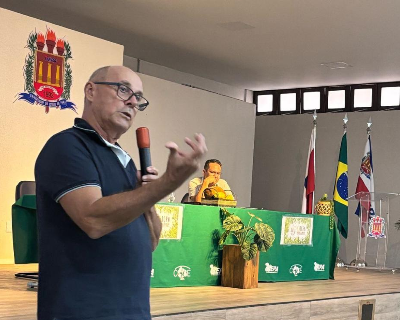 Homem careca de óculos e polo azul fala ao microfone em um palco. Ao fundo, mesa com toalha verde e outro homem sentado. Na parede, brasão da UEPA. À direita, bandeiras do Pará, Brasil e Belém. Ambiente de auditório.