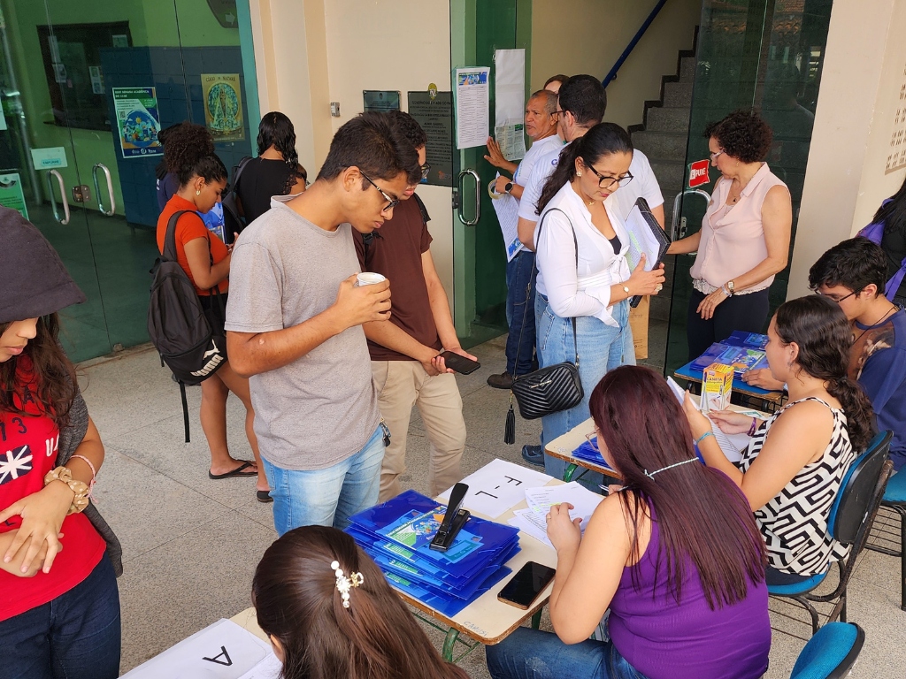 A foto mostra um grupo de pessoas em frente a uma entrada de prédio, onde há mesas organizadas para credenciamento ou inscrição. Algumas pessoas estão sentadas, preenchendo papéis e conferindo listas, enquanto outras aguardam em pé, segurando documentos ou conversando. O ambiente é movimentado, com estudantes e adultos participando da atividade.