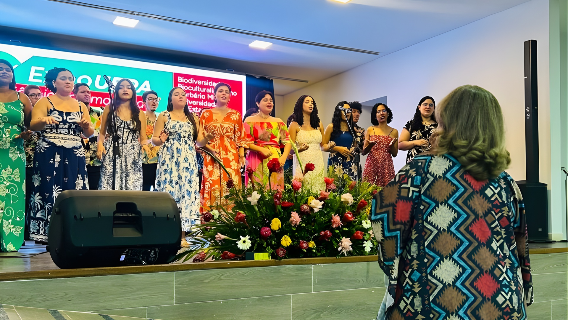 Um grupo de cerca de quinze jovens canta em um palco iluminado, usando vestidos coloridos com estampas florais ou inspiradas na cultura amazônica. Eles estão alinhados atrás de microfones, com um grande arranjo de flores tropicais à frente. No canto inferior direito, de costas, aparece uma mulher de cabelos claros e curtos observando a apresentação; ela veste uma blusa com padrão geométrico. Ao fundo, um telão exibe textos sobre biodiversidade e cultura.