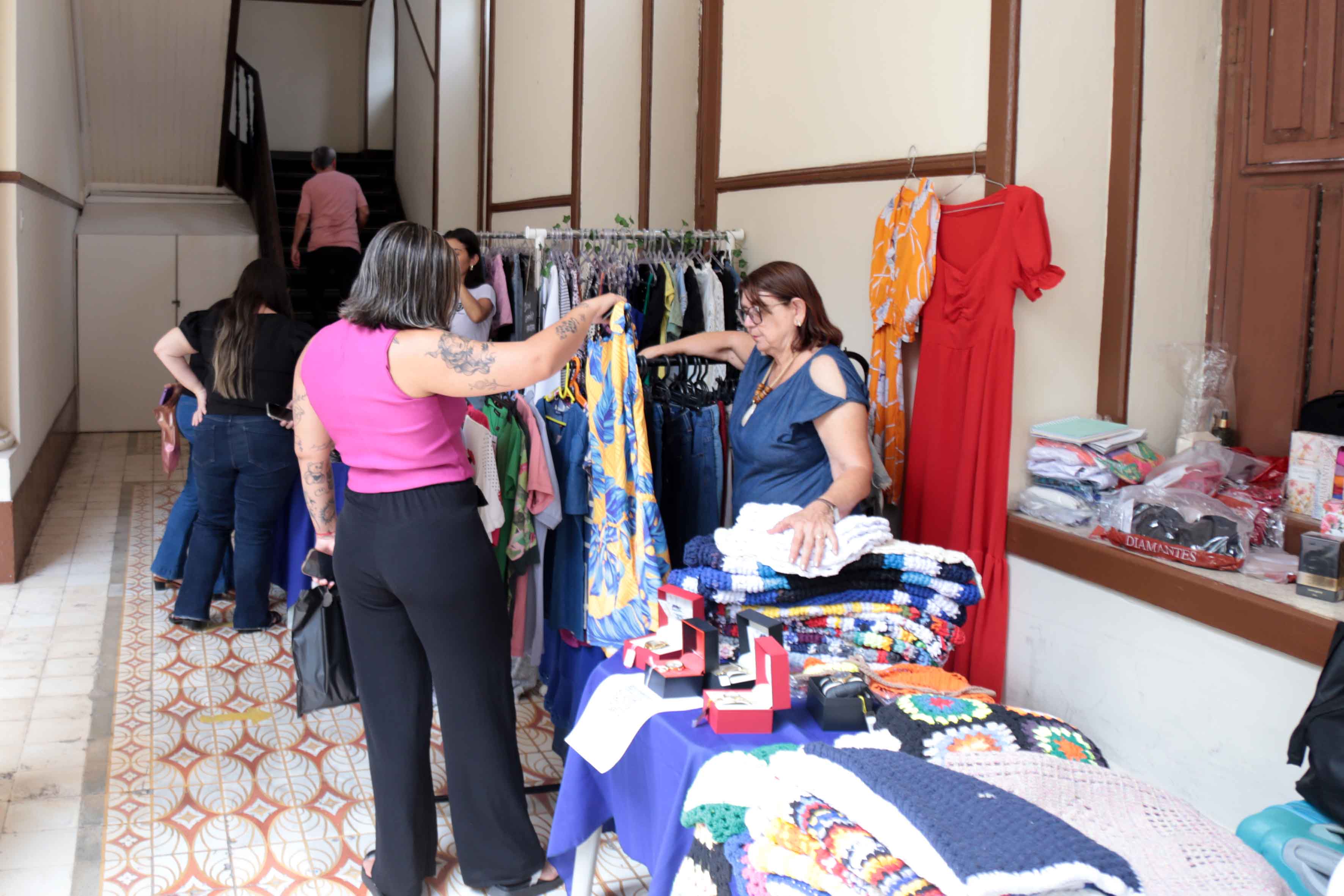 A imagem mostra uma feira em ambiente interno, onde duas mulheres interagem em um estande de roupas e produtos artesanais. Uma delas observa uma peça de vestuário em um cabide, enquanto a outra organiza itens sobre a mesa, que inclui peças de crochê, acessórios e caixas. A