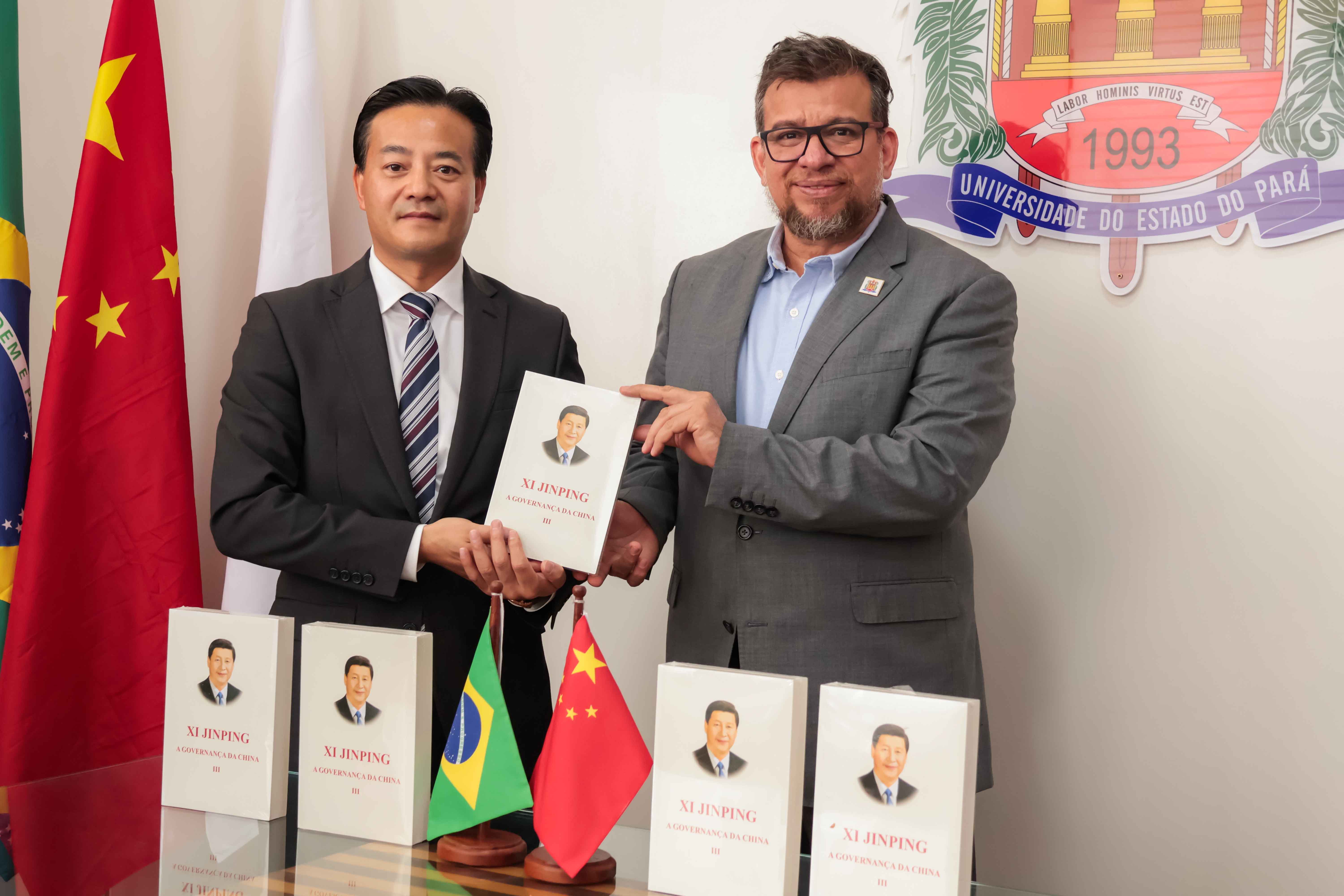 A imagem mostra duas pessoas lado a lado segurando um livro intitulado "XI JINPING: A GOVERNANÇA DA CHINA III". À esquerda, um homem de terno escuro e gravata está posicionado em frente a uma bandeira da China e, à direita, um homem de terno cinza e camisa azul, com óculos, está em frente ao brasão da Universidade do Estado do Pará (Uepa). Sobre a mesa à frente deles, estão pequenas bandeiras do Brasil e da China, além de outros exemplares do mesmo livro.