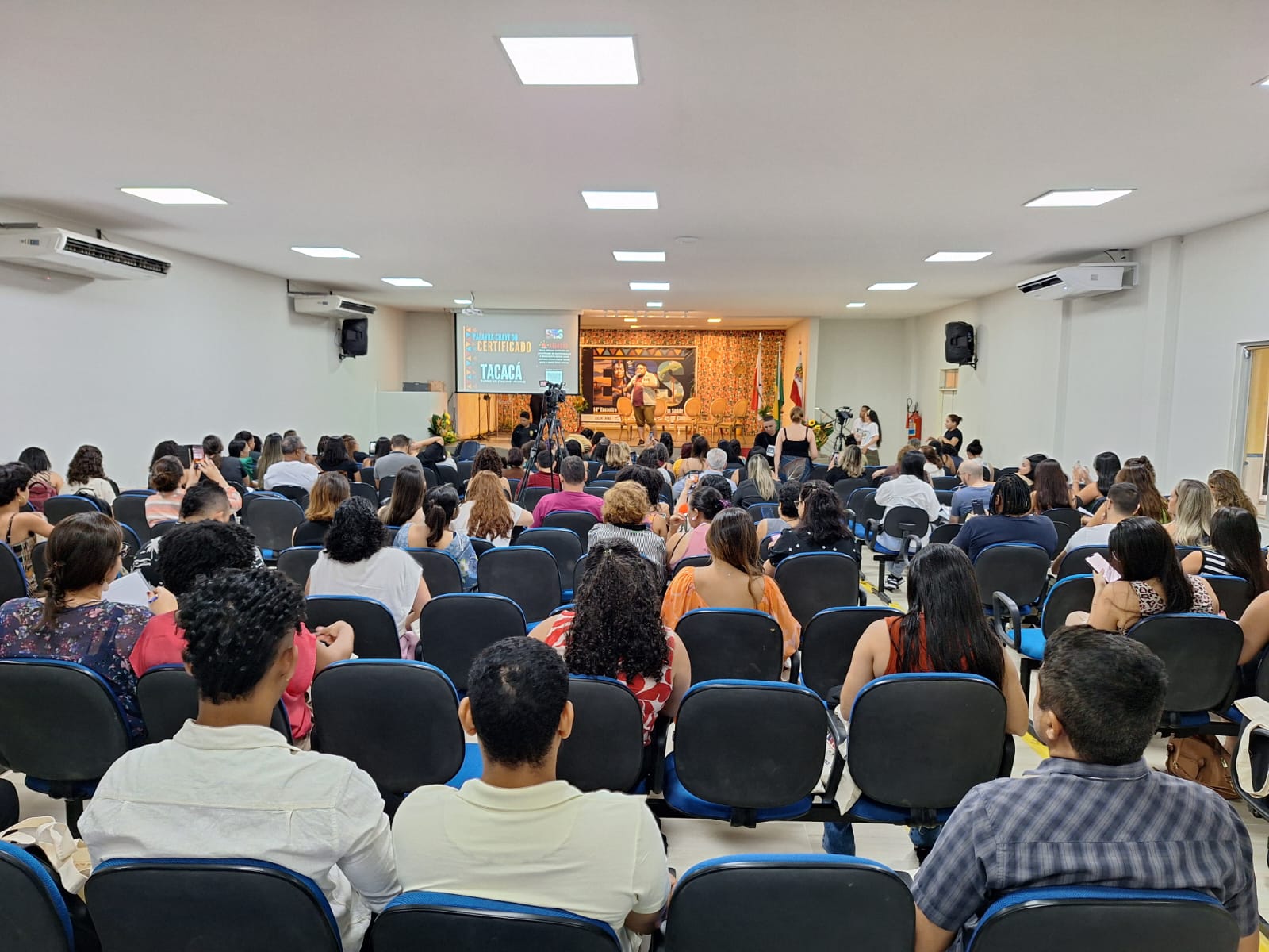 A imagem mostra um evento em um auditório, com várias pessoas sentadas em cadeiras voltadas para um palco. No palco, há indivíduos em pé na frente de um banner com texto, e uma tela exibindo informações adicionais. O ambiente é de uma conferência com um momento de interesse coletivo para os participantes.