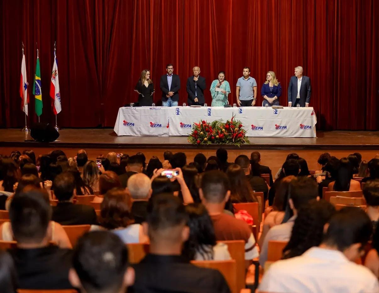 A imagem mostra várias pessoas sentadas, assistindo à cerimônia no palco. No palco, há uma mesa longa e gestores estão de pé, atrás dela. 