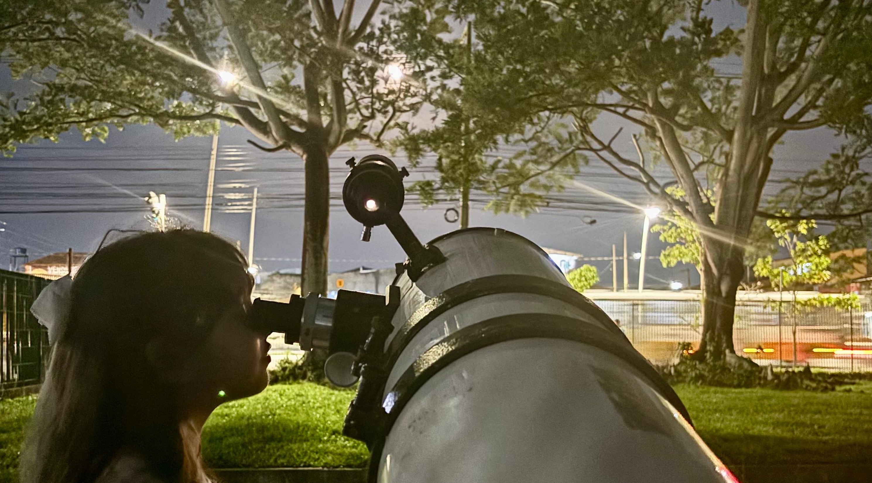 A imagem mostra uma jovem observando o céu noturno através de um grande telescópio. A cena se passa na área externa do Centro de Ciências e Planetário do Pará (CCPPA), um ambiente cercado por árvores, com luzes urbanas e fios elétricos ao fundo. O céu escuro e parcialmente iluminado pela lua e postes de luz, destaca a silhueta da menina e do telescópio.