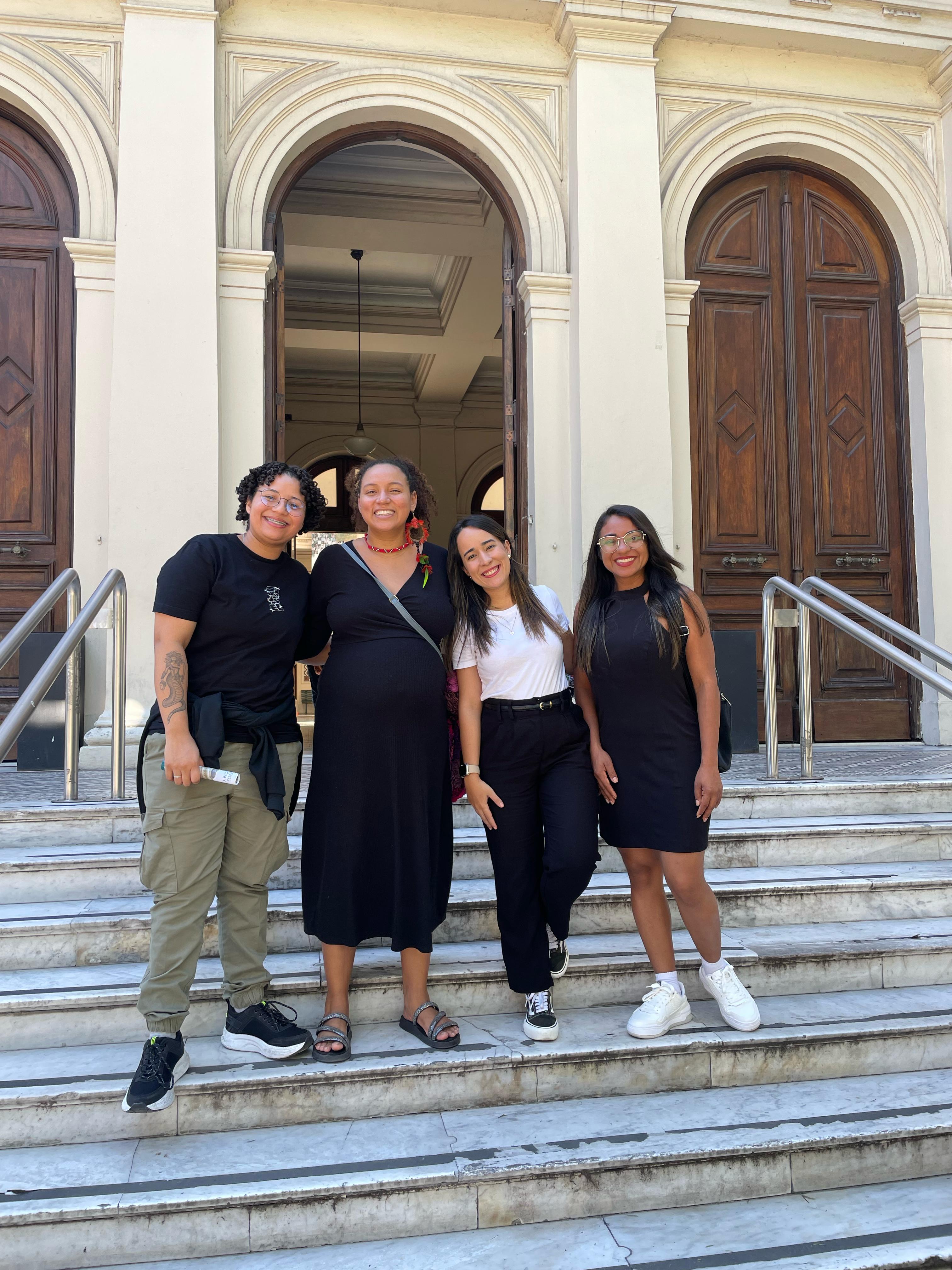 A imagem mostra grupo de quatro pessoas em frente a um prédio histórico, com portas e arcos altos de madeira.  Elas posam sorrindo nos degraus da entrada do prédio.