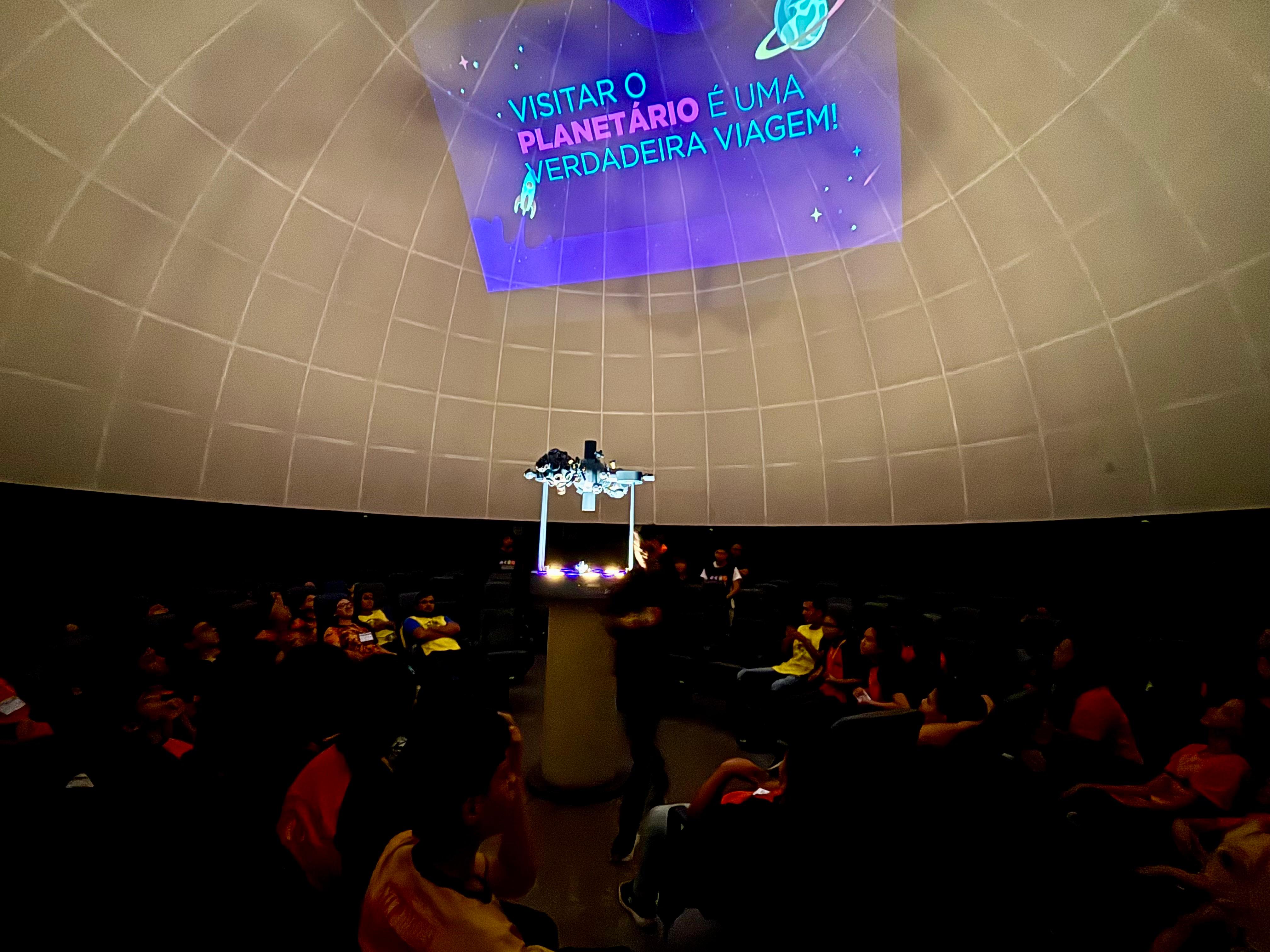 A imagem mostra o interior da cúpula de projeção Kwarahy. No centro, há um projetor planetário, utilizado para simular o céu. Em volta dele, crianças e adolescentes estão sentados em poltronas, olhando para cima, atentos à projeção.
