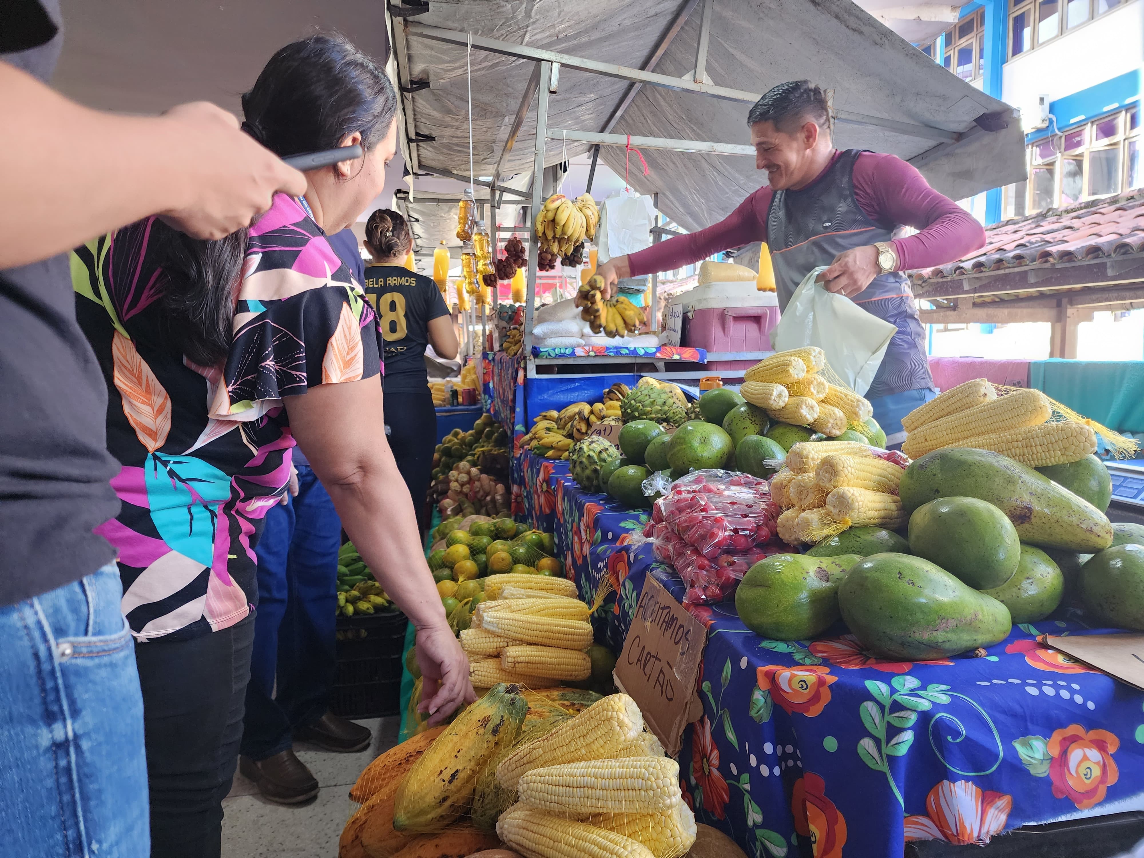 Em uma feira da agricultura familiar, um homem feirante sorri enquanto entrega um cacho de bananas a uma mulher que escolhe produtos expostos sobre uma toalha colorida. Há espigas de milho, mangas, cacaus, abacates e pacotes de frutas vermelhas organizados na barraca. Ao fundo, outras pessoas circulam entre as bancas, sob lonas que cobrem a estrutura da feira. Um cartaz avisa que o local aceita cartão.