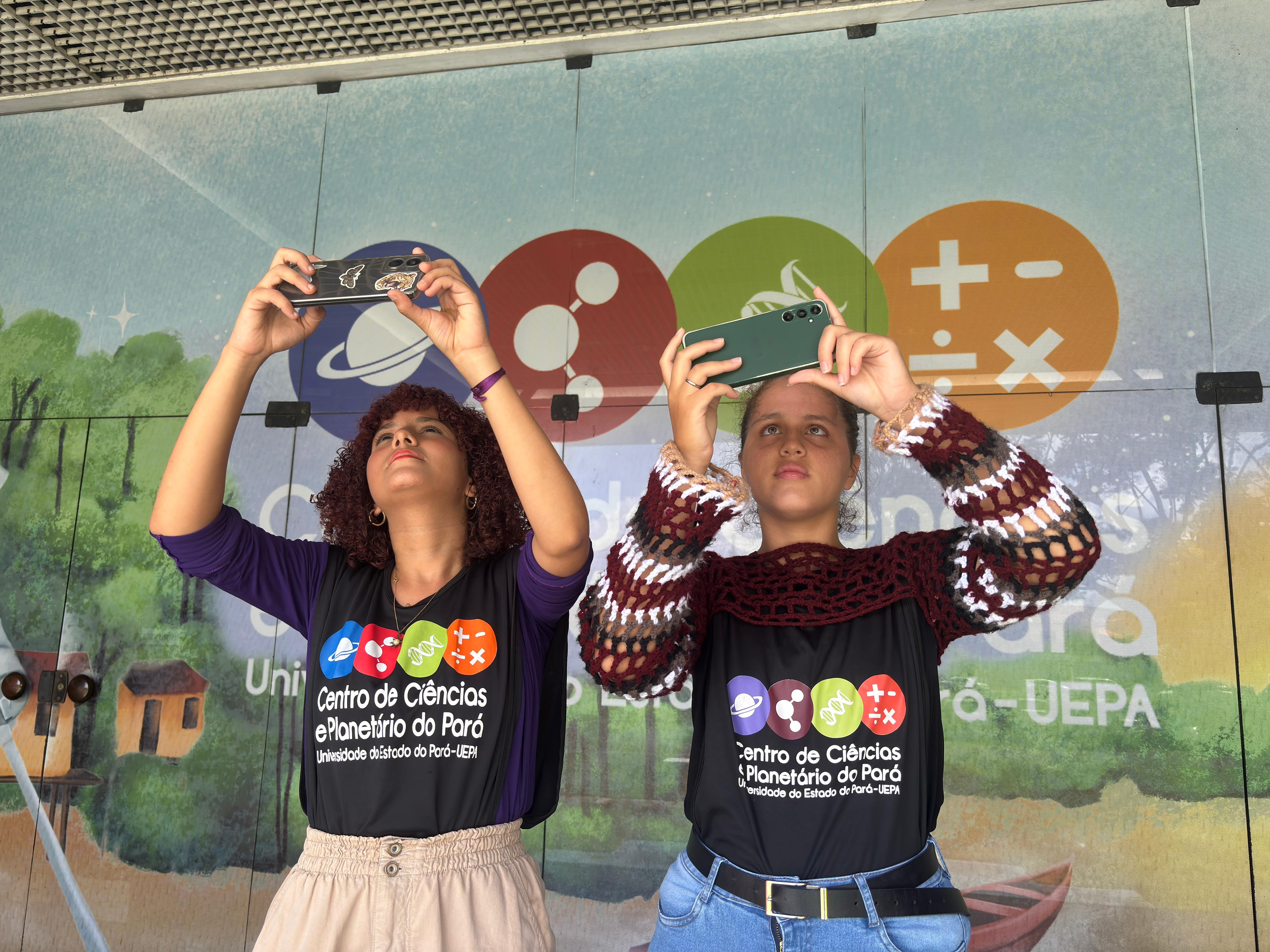 A imagem mostra duas estagiárias do CCPPA, segurando celulares em posição elevada, fazendo fotos. A modalidade de astrofotografia do VIII Concurso Ciência e Arte permite a participação dos inscritos com o registro de imagens através do aparelho celular.