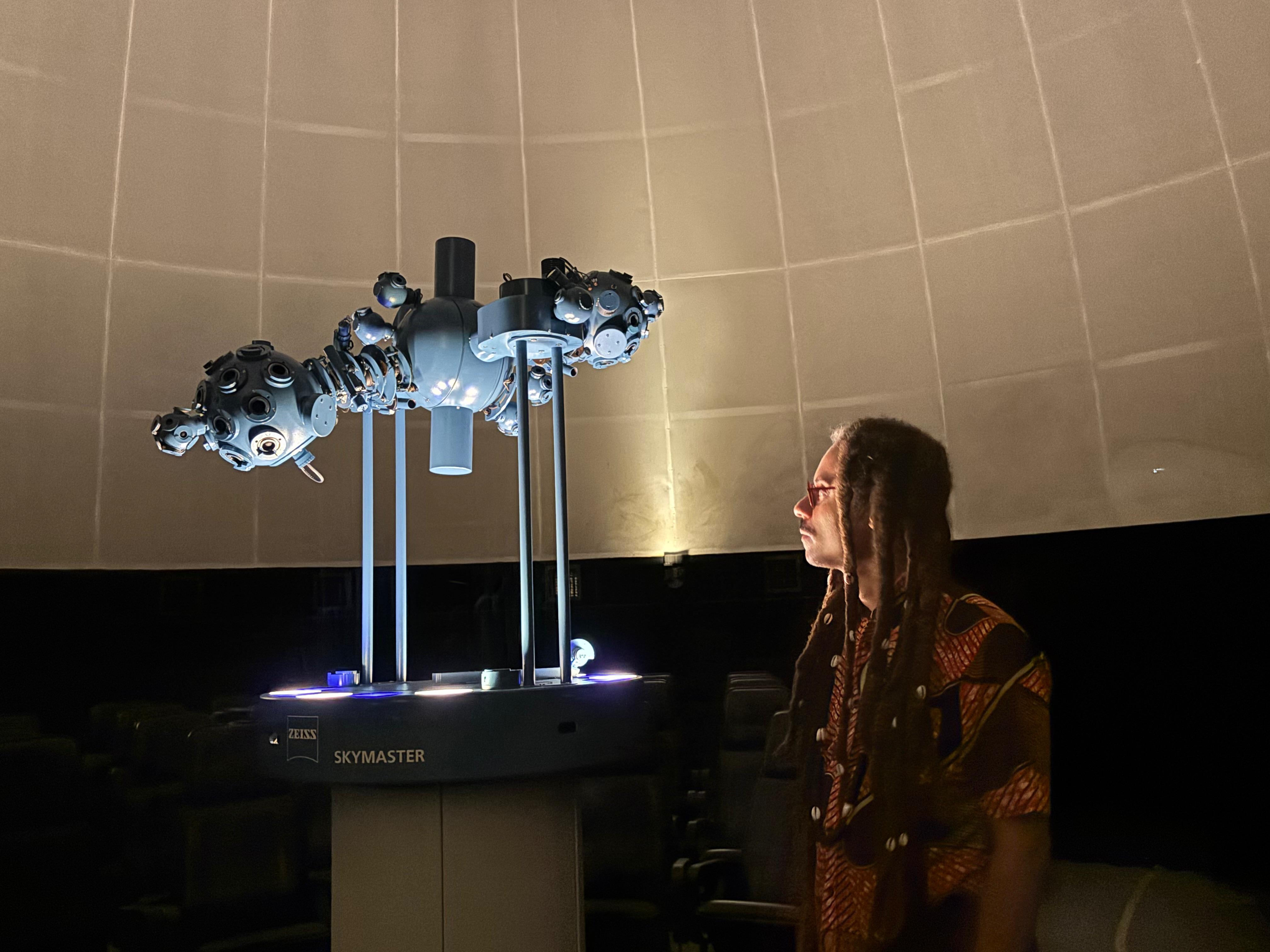 A imagem mostra o interior da cúpula Kwarahy, do CCPPA, com iluminação suave e atmosfera contemplativa. Em destaque no centro, está um projetor planetário ZEISS modelo Skymaster, equipamento usado para simular o céu estrelado no teto do domo. À direita da imagem, observa-se o artista visual Rafael RG.