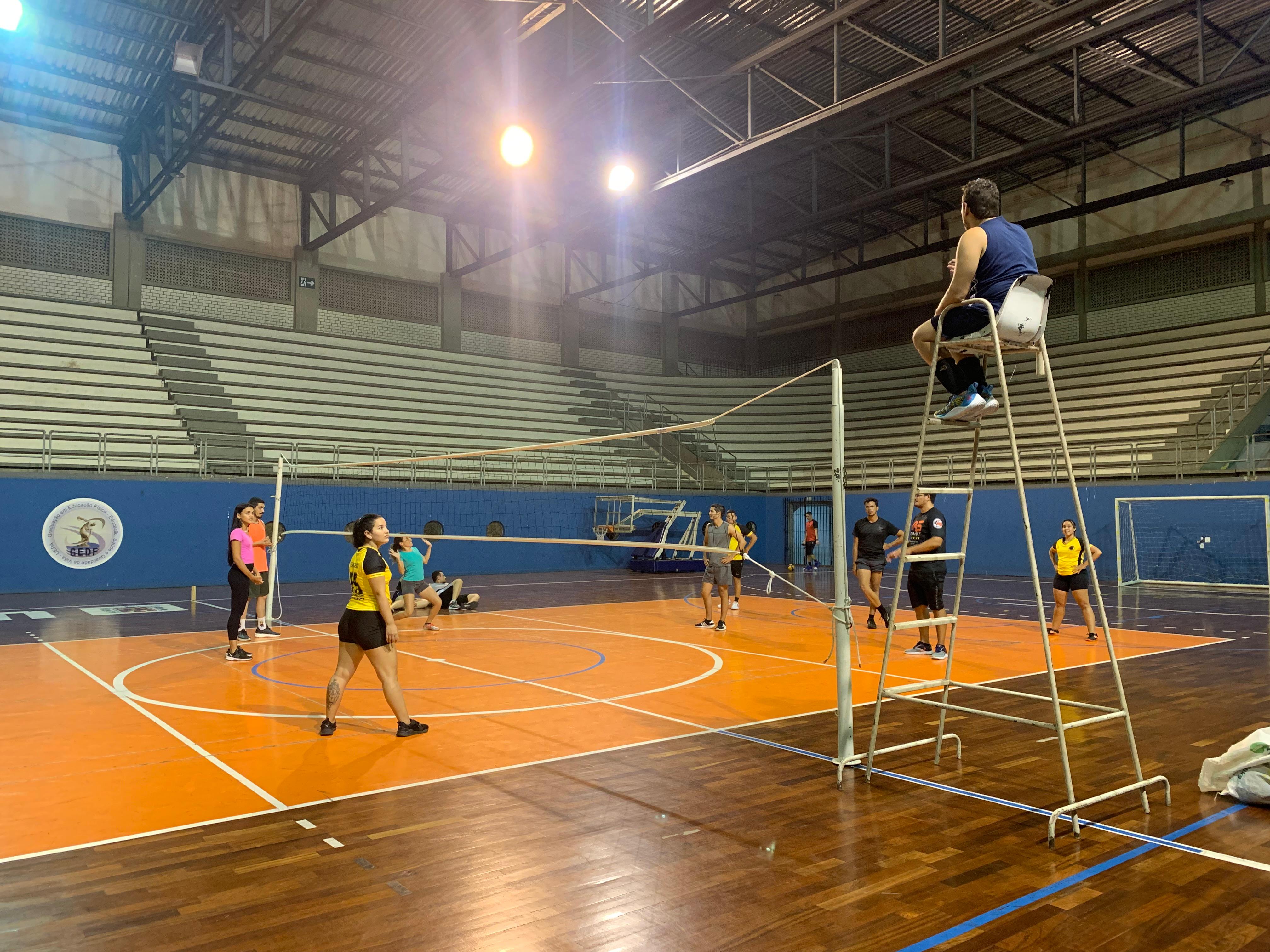 A imagem mostra um jogo de vôlei em um ginásio poliesportivo, com árbitro na cadeira elevada, jogadores em quadra com piso de madeira, arquibancadas vazias ao fundo. e logotipo ligado à área de educação física.