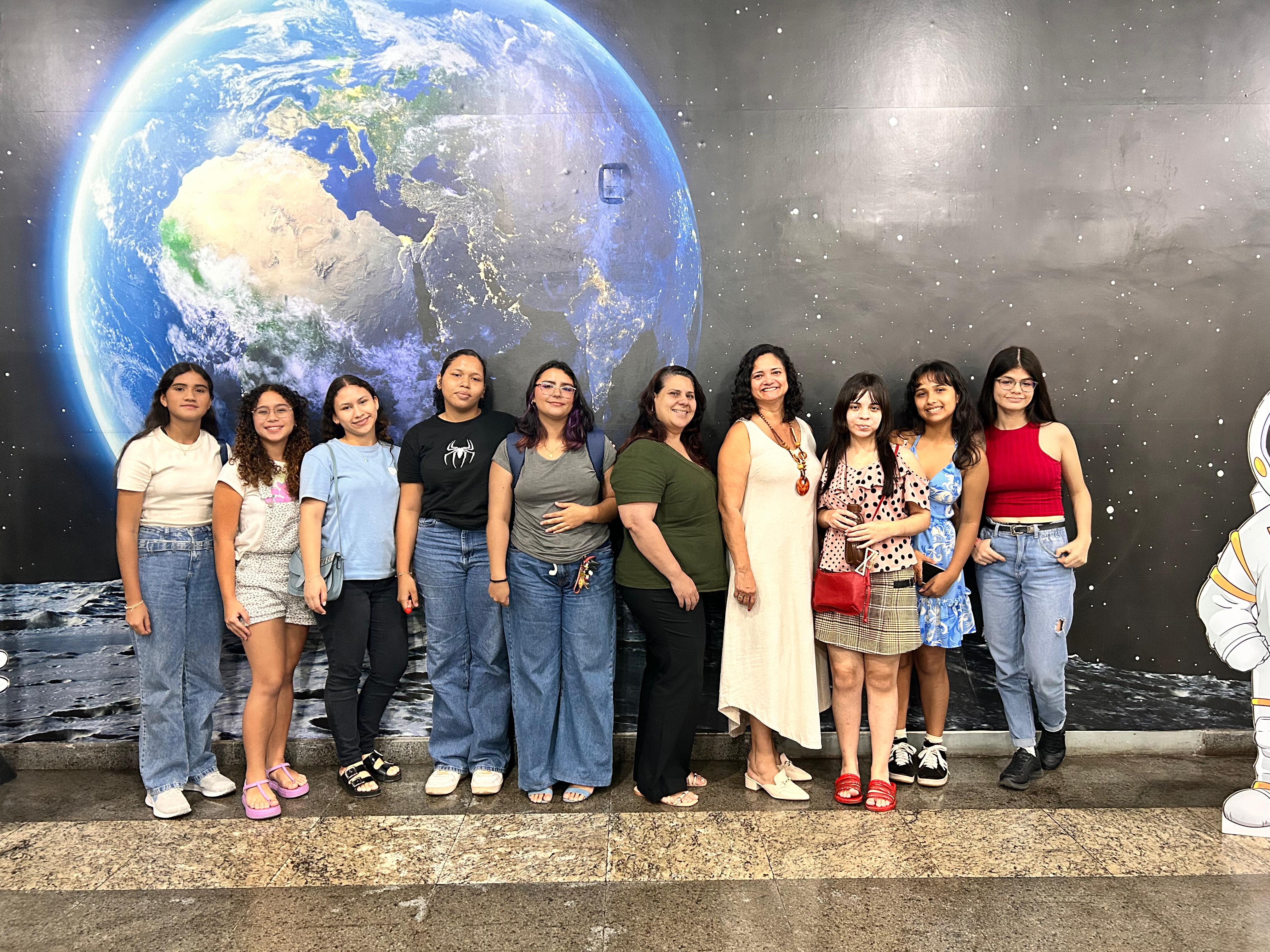 A imagem mostra as meninas que integram o projeto Astrociência, junto a professoras do CCPPA, posando para uma foto em frente a um mural espacial. Ao fundo, há uma imagem grande do planeta Terra vista do espaço, com detalhes realistas, e o chão simula a superfície da Lua.