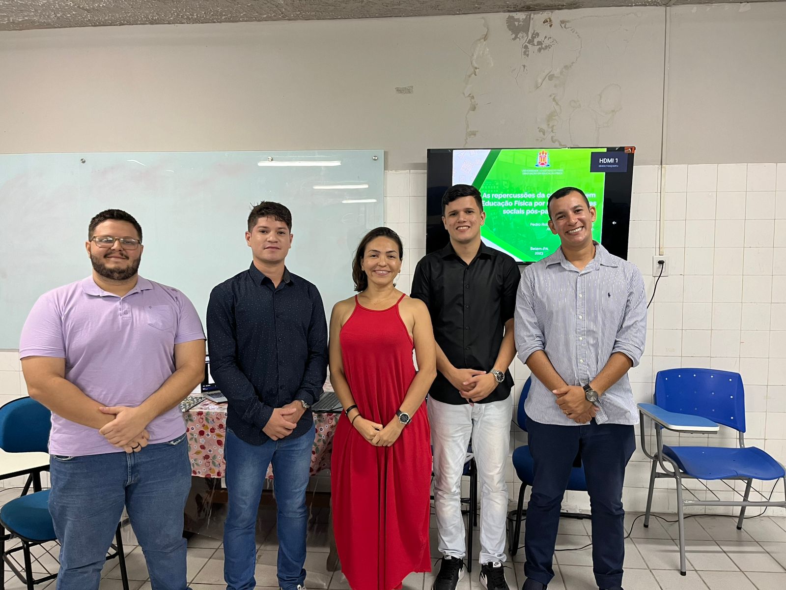 A imagem mostra cinco pessoas em pé, sorrindo lado a lado, posando para a foto em uma sala de aula. Atrás delas, há uma tela ligada exibindo uma apresentação com o título “As repercussões da atuação em Educação Física por meio das redes sociais pós-pandemia”. O ambiente tem um quadro branco, paredes de azulejo e algumas cadeiras.