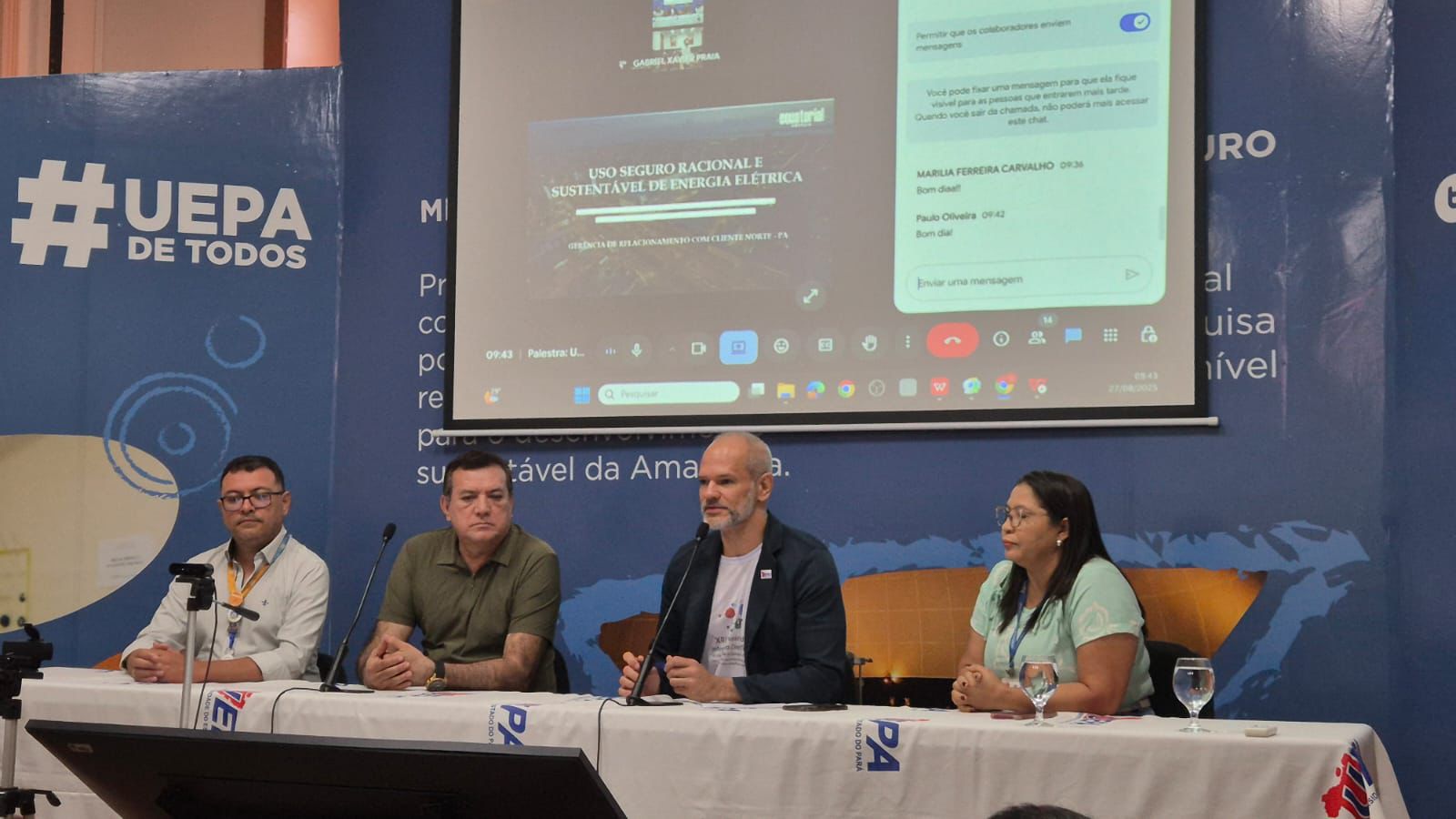 Na foto, quatro pessoas estão sentadas à mesa de um evento institucional da Uepa, participando de uma mesa-redonda sobre o uso sustentável de energia elétrica. Atrás deles, um telão exibe a apresentação e a transmissão online.