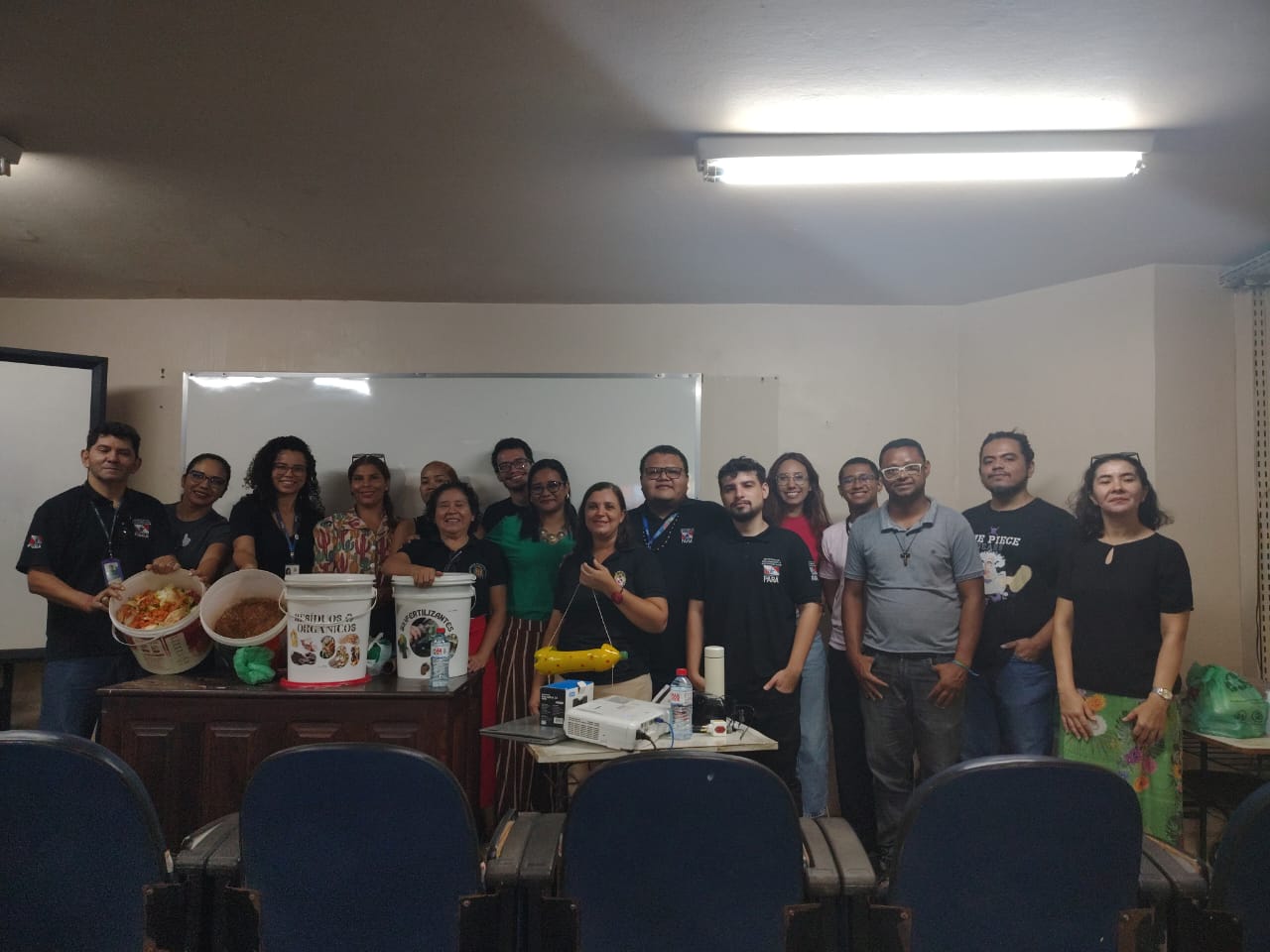 Na foto, um grupo de pessoas posa reunido em uma sala de aula ao final de uma atividade prática de compostagem. Algumas seguram baldes com resíduos orgânicos e materiais recicláveis, simbolizando as ações de sustentabilidade discutidas no encontro.