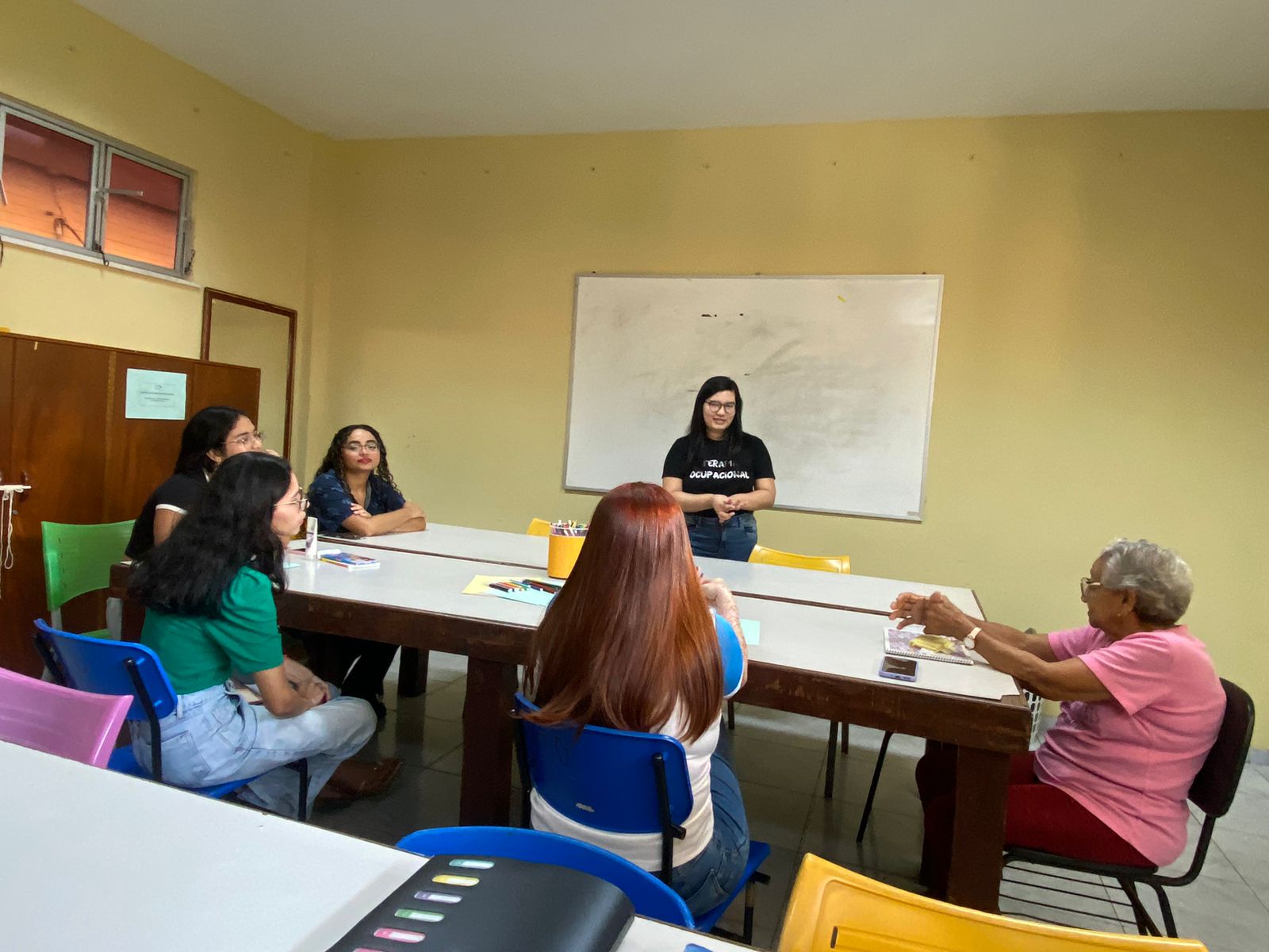Pessoas reunidas em uma sala, sentadas em uma mesa, os participantes realizam atividades como colorir, desenhar, com materiais de arte espalhados sobre a mesa.