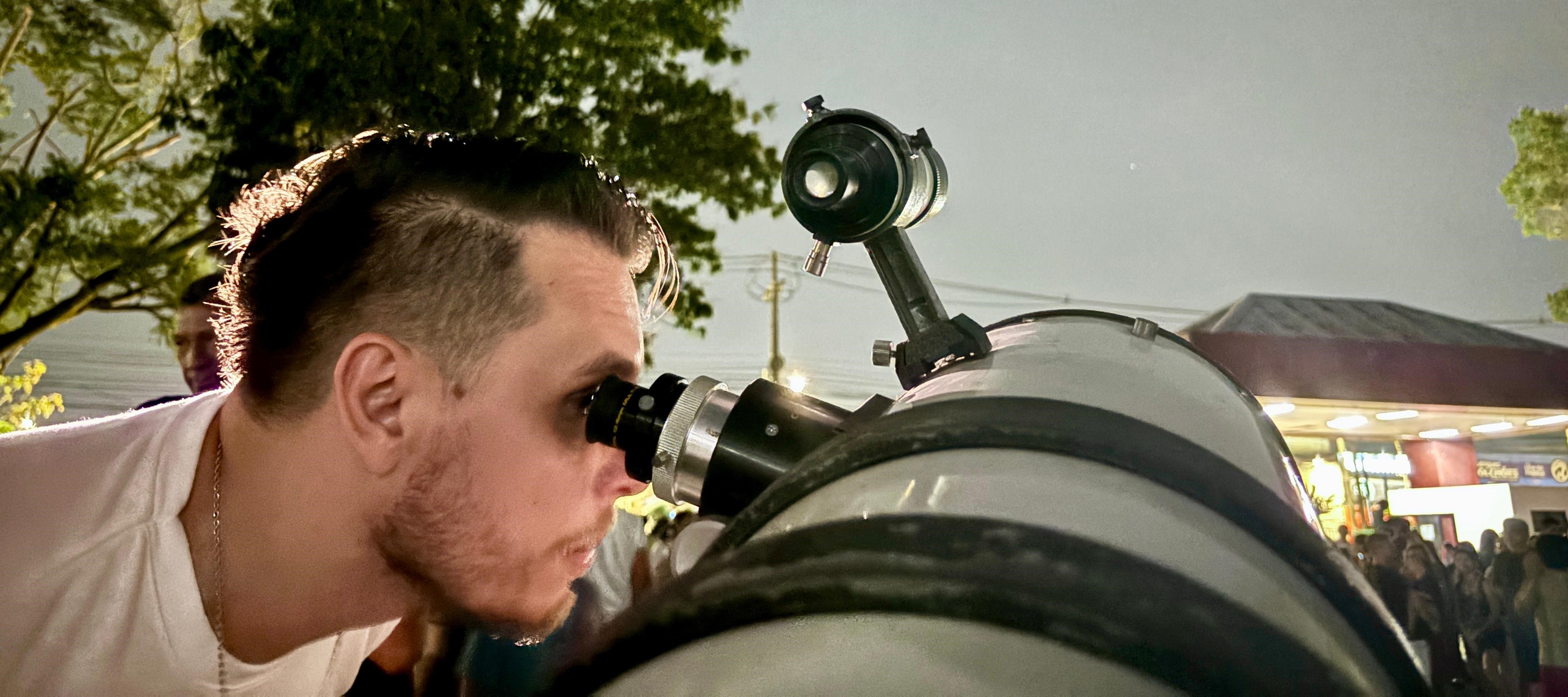 A imagem mostra uma pessoa observando o céu noturno através de um telescópio. A cena acontece na área externa do Centro de Ciências e Planetário do Pará. O clima transmite curiosidade científica e encantamento com o Universo.