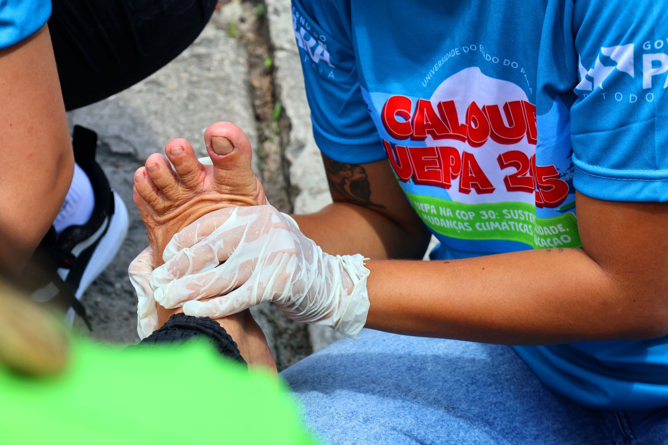 A imagem mostra uma pessoa fazendo atendimento e oferecendo cuidados em um pé de um romeiro