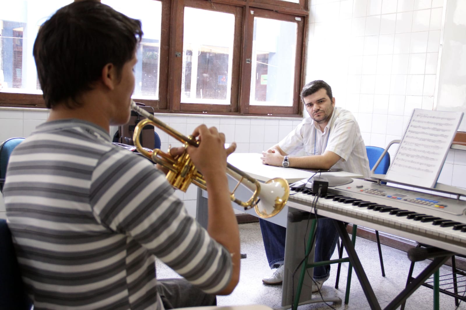 Um jovem toca um trompete dourado enquanto um professor  de camisa branca e calça jeans, o observa atentamente em uma sala de aula com janelas de madeira. Um teclado musical e partituras estão sobre a mesa à frente.
