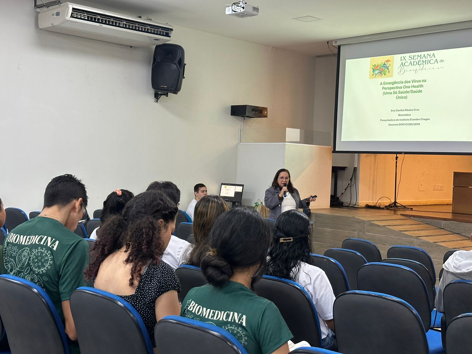 A imagem mostra uma palestra em andamento, apresentando para uma plateia de alunos em um auditório