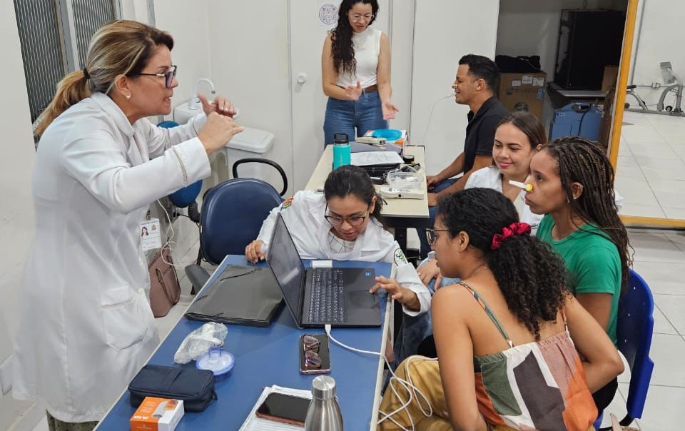 A imagem mostra um grupo em treinamento prático dentro de uma sala. Uma instrutora de jaleco orienta os participantes reunidos ao redor de uma mesa com um laptop, todos focados na atividade..