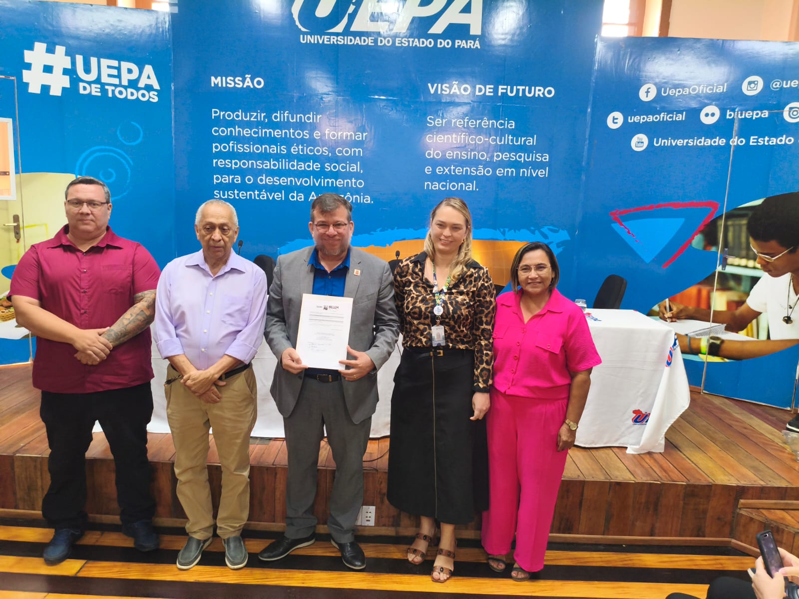 A imagem mostra um grupo de pessoas em pé em um evento institucional e no centro, um dos membros segura um documento oficial.