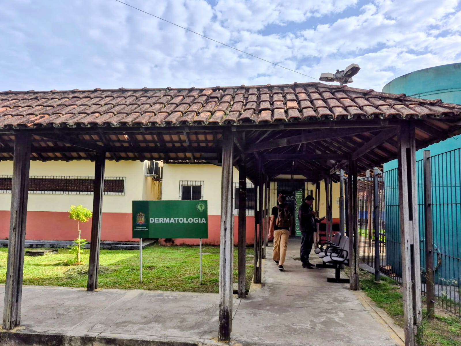 A imagem mostra um corredor coberto em frente a uma instituição de saúde, com pessoas andando e uma placa onde está escrito: Dermatologia. Duas pessoas estão andando pelo corredor.