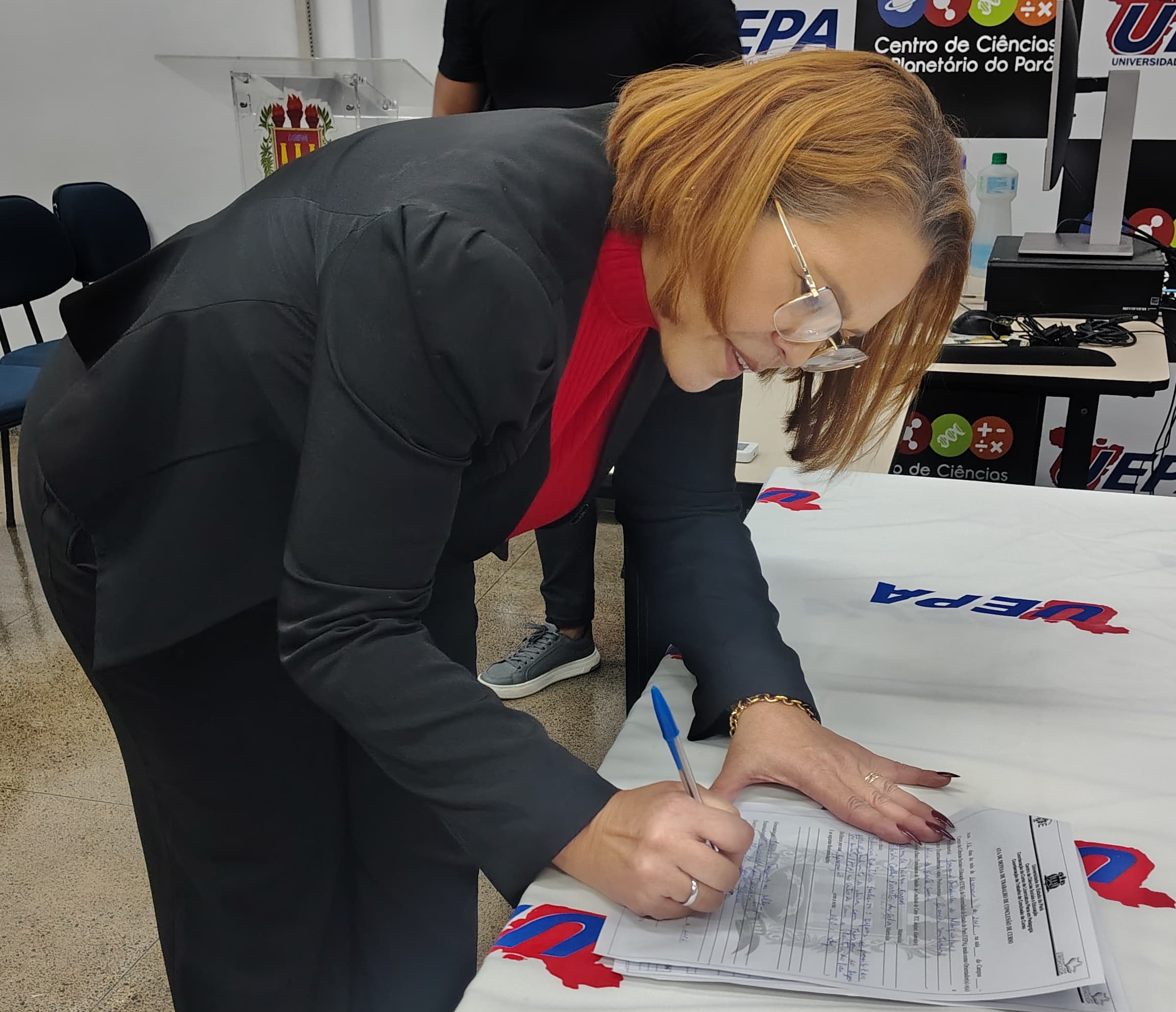 A imagem mostra Camila Barros, sobre uma mesa enquanto assina um documento. Ela usa blazer preto, blusa vermelha e óculos de armação transparente. Seu cabelo é curto, liso e em tom loiro-acobreado. Na mão direita, ela segura uma caneta azul e escreve sobre um termo oficial.