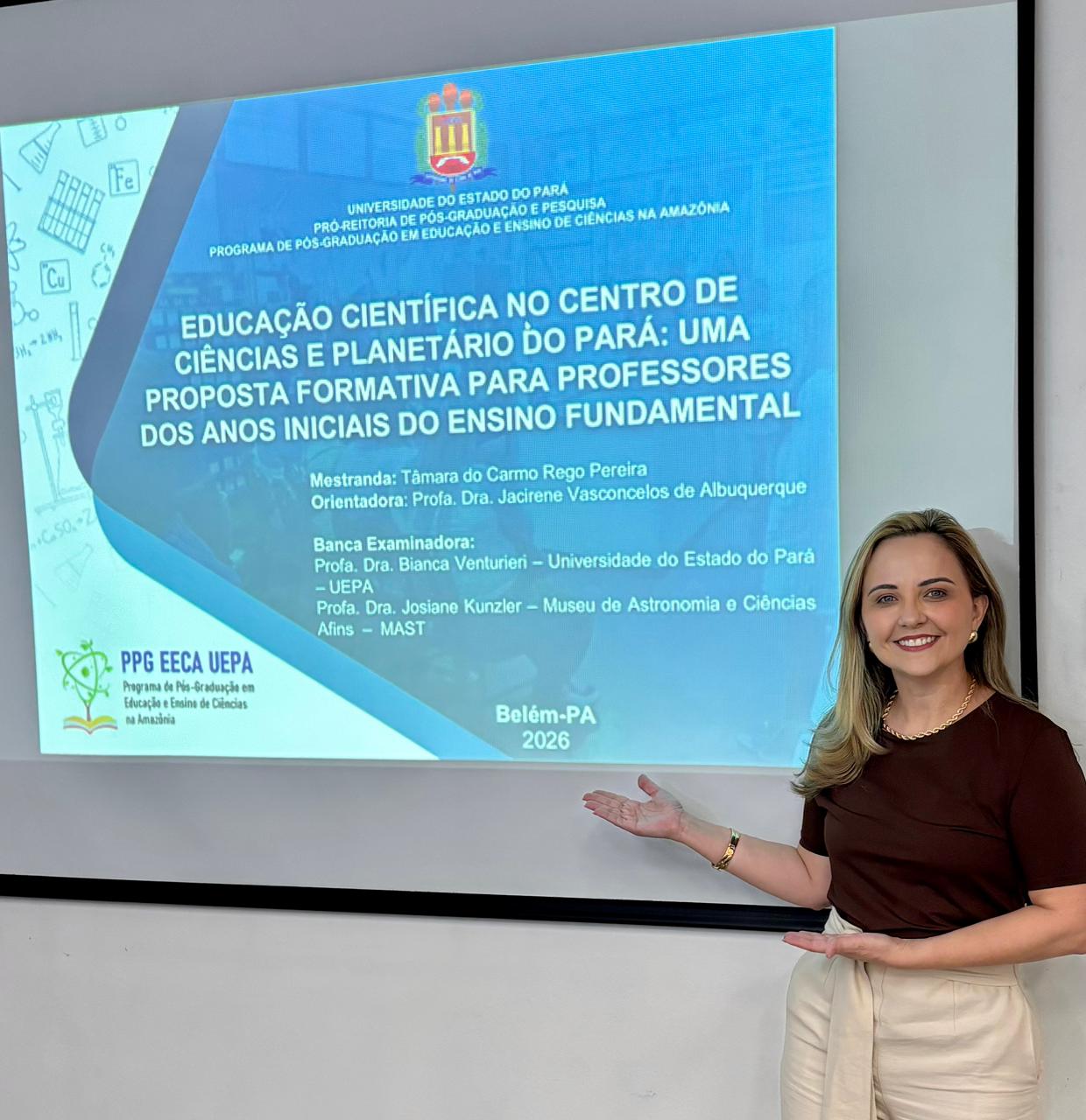 A imagem mostra Tâmara Pereira em pé ao lado de uma tela de projeção, sorrindo e apontando com as mãos abertas para o slide exibido durante a apresentação da sua dissertação. Ela tem cabelos loiros, usa uma blusa marrom de manga curta e uma calça clara.