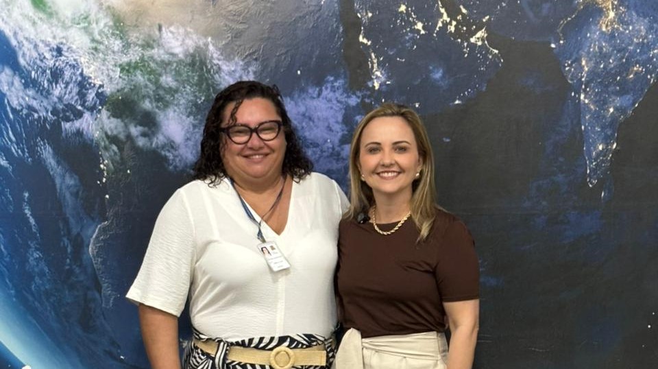 A foto mostra Jacirene Albuquerque e Tâmara Pereira em pé, lado a lado, sorrindo para a câmera. Elas estão diante de um grande painel com uma imagem da Terra vista do espaço, que compõe o hall do Centro de Ciências e Planetário do Pará.