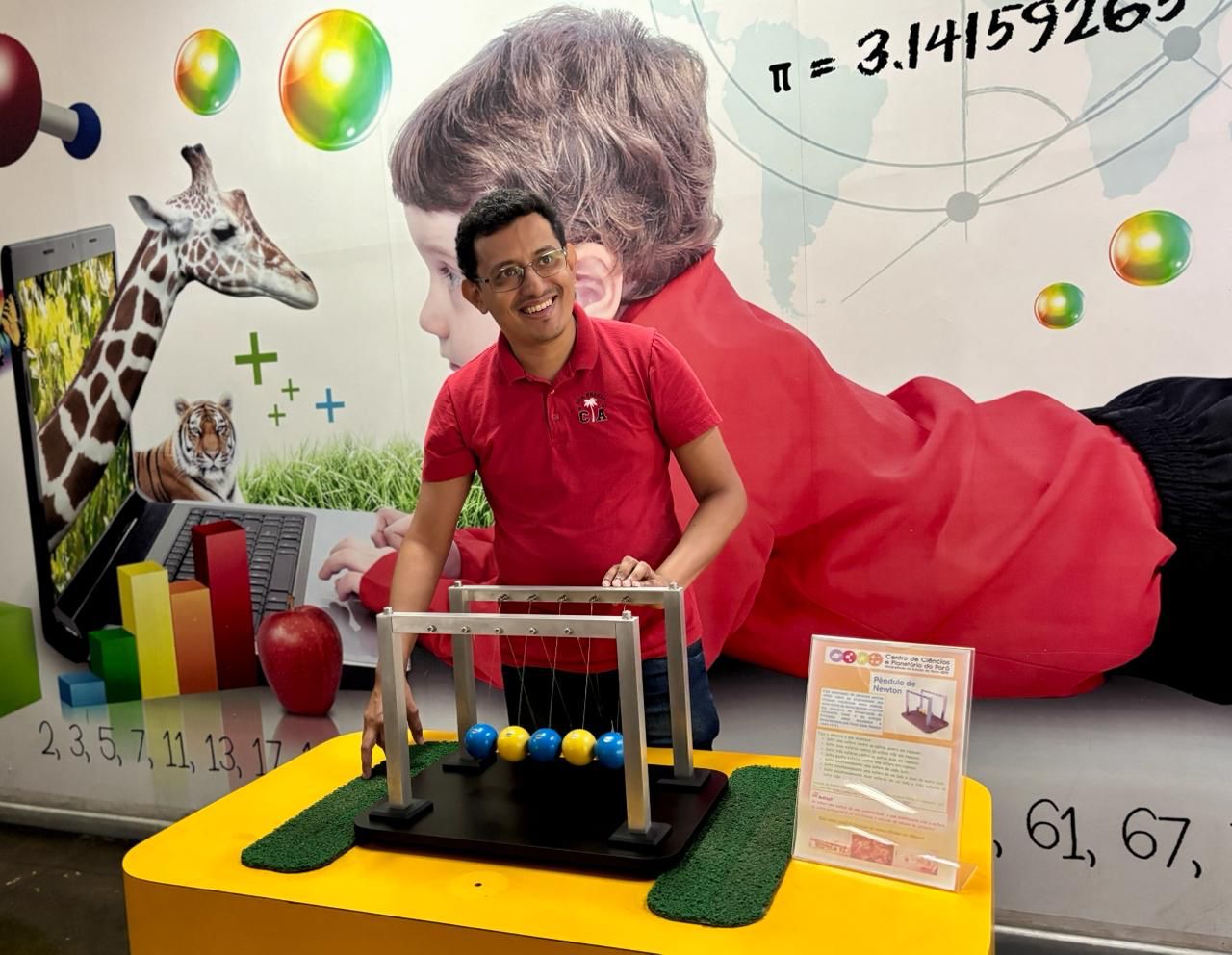 A imagem mostra o docente da Uepa, Reginaldo Corrêa, posicionado atrás de uma mesa amarela no Centro de Ciências. Sobre a mesa há um experimento conhecido como pêndulo de Newton, formado por uma estrutura metálica com cinco esferas suspensas por fios.
