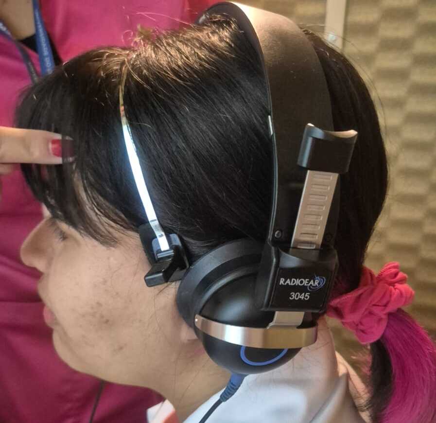 A imagem mostra uma pessoa jovem usando fones de ouvido grandes, aparentemente passando por um teste ou avaliação auditiva