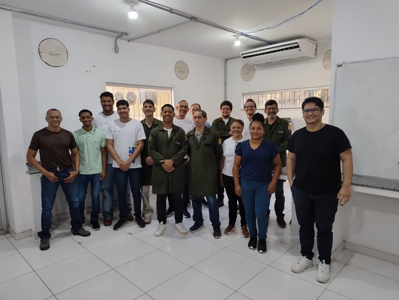 Um grupo diversificado de pessoas dentro de uma sala de aula ou laboratório, sugerindo um ambiente de aprendizado ou trabalho. As pessoas estão em pé e em fileiras, olhando para a câmera. 