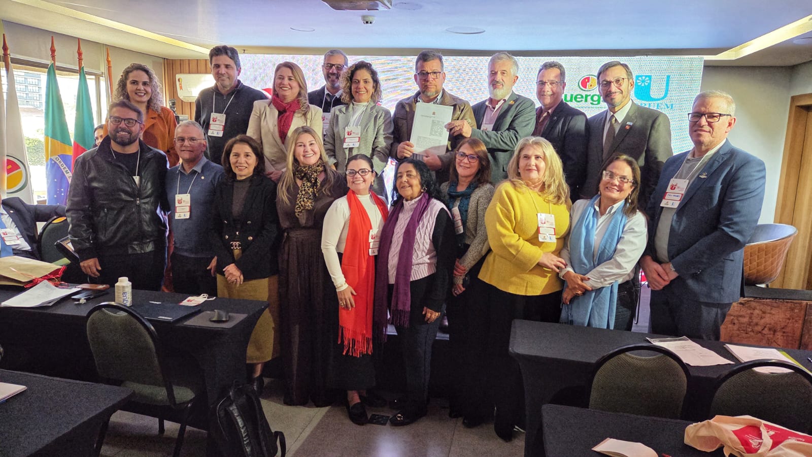 Foto de grupo em ambiente interno de evento institucional. Ao centro, cerca de 17 pessoas posam para a câmera, entre homens e mulheres de diferentes idades, usando crachás de identificação. Algumas estão em pé e outras levemente à frente, formando duas fileiras. No centro da imagem, um homem segura um documento enquanto outro participante aponta para ele. Ao fundo, há bandeiras e um painel iluminado com logotipos de universidades e instituições.