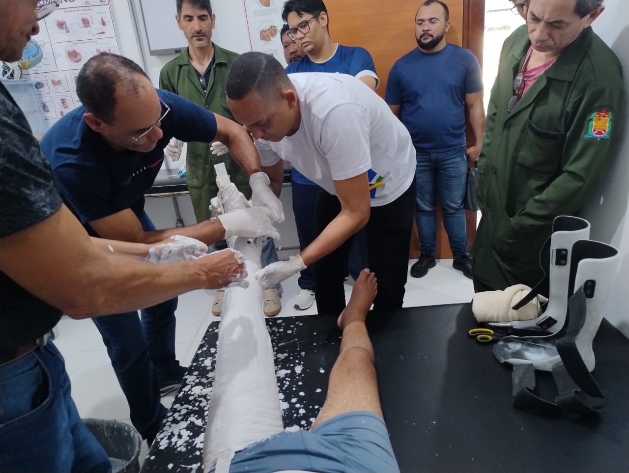 Um grupo de pessoas, alguns com jalecos, estão focados em aplicar um gesso em uma pessoa, simulando um cenário de aprendizado em um ambiente como um laboratório. Algumas pessoas com luvas moldam o gesso em uma perna, enquanto outras observam atentamente.