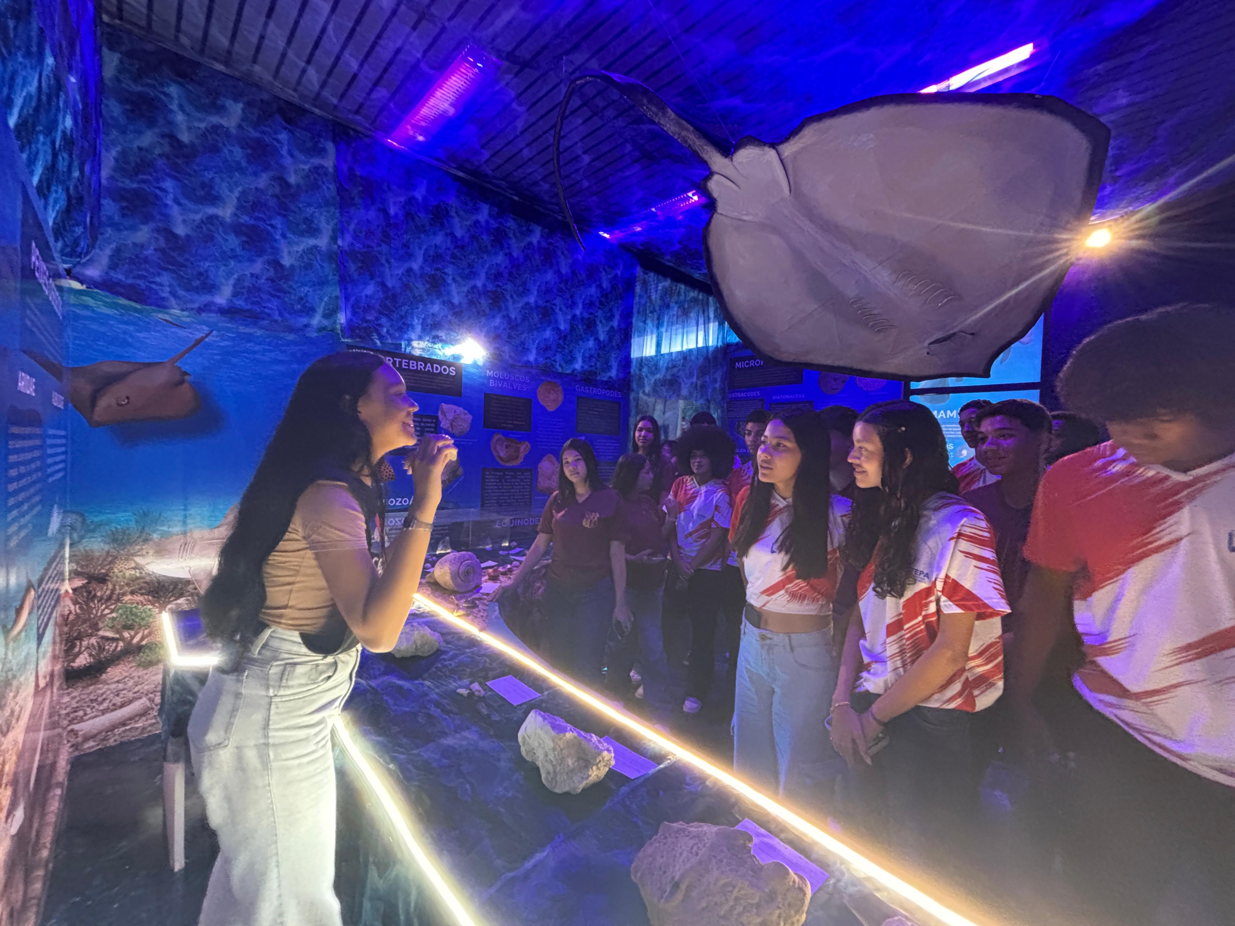 A imagem mostra uma visita guiada à exposição “O Mar Perdido de Pirabas”, realizada no Centro de Ciências e Planetário do Pará (CCPPA). Em primeiro plano, uma mediadora apresenta informações aos visitantes enquanto aponta para peças expostas em uma vitrine iluminada. Ao redor, um grupo de estudantes acompanha atentamente as explicações.