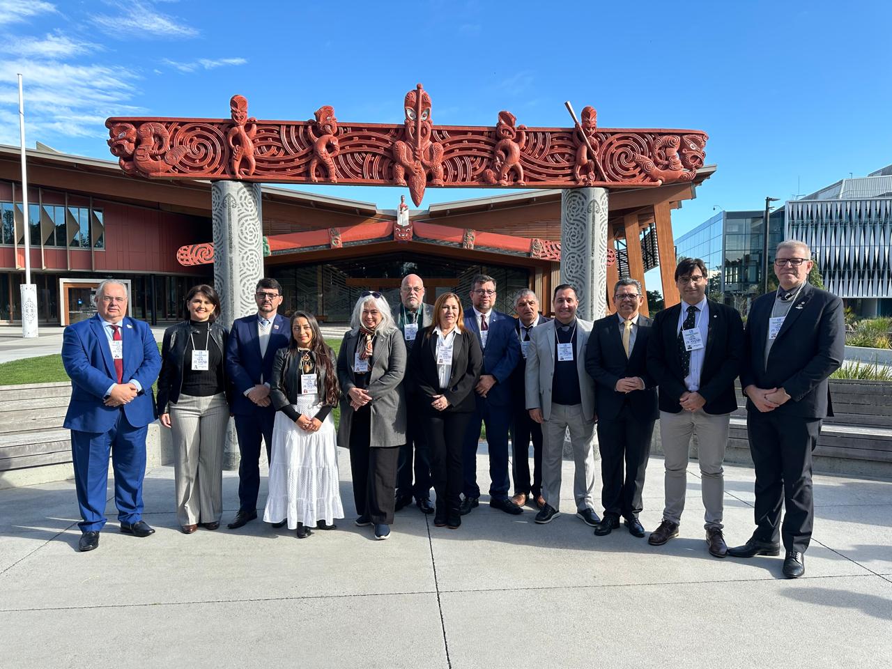 A imagem mostra um grupo de 15 pessoas posando para uma foto em frente a uma construção tradicional maori, localizada na Universidade de Waikato, na Nova Zelândia. O grupo está alinhado e veste trajes formais, indicando uma visita institucional ou missão internacional. Ao fundo, é possível ver detalhes arquitetônicos típicos da cultura maori, com entalhes em madeira vermelha e pilares esculpidos. O céu está azul, e o clima é ensolarado.