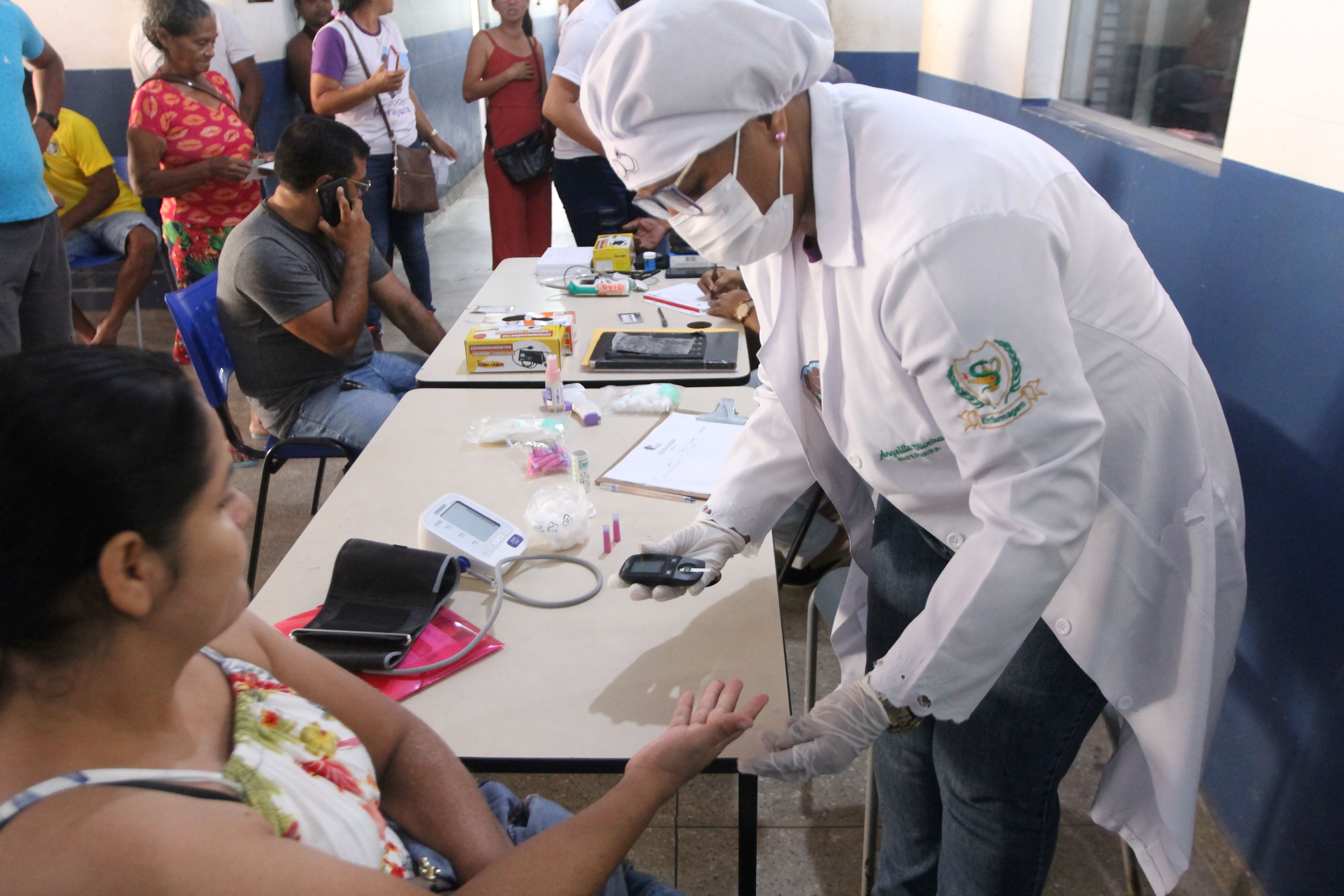 A imagem mostra um ambiente interno onde um profissional de saúde, vestindo um jaleco branco com um emblema, luvas e um gorro, está atendendo pessoas sentadas em mesas. O profissional parece estar preparando ou administrando algum tipo de procedimento ou teste médico. Há várias pessoas ao fundo que parecem estar esperando sua vez ou observando o processo. Na mesa, há diversos itens, incluindo o que parece ser equipamento médico, como um monitor de pressão arterial e possivelmente alguns recipientes.