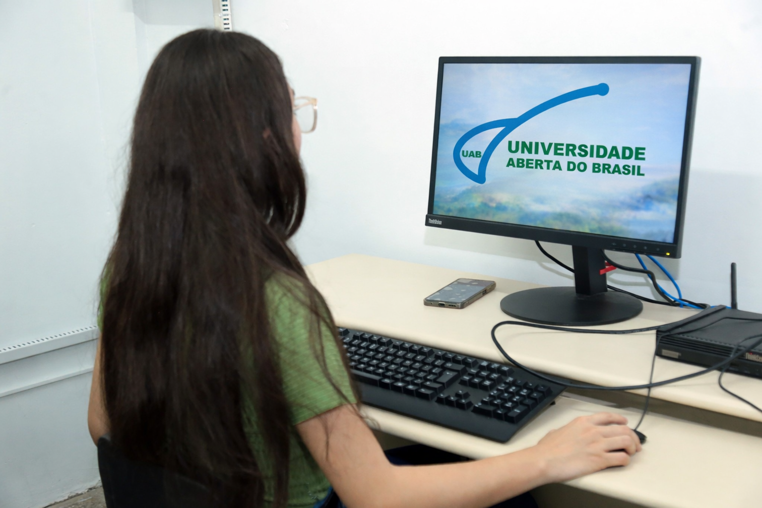 A imagem mostra uma estudante sentada em uma estação de trabalho de informática, olhando para o monitor de um computador que exibe a marca da Universidade Aberta do Brasil (UAB). Ela está em um ambiente claro e organizado, com teclado, mouse e outros equipamentos sobre a mesa e transmite a ideia de ensino a distância, acesso à tecnologia e apoio institucional à formação acadêmica.