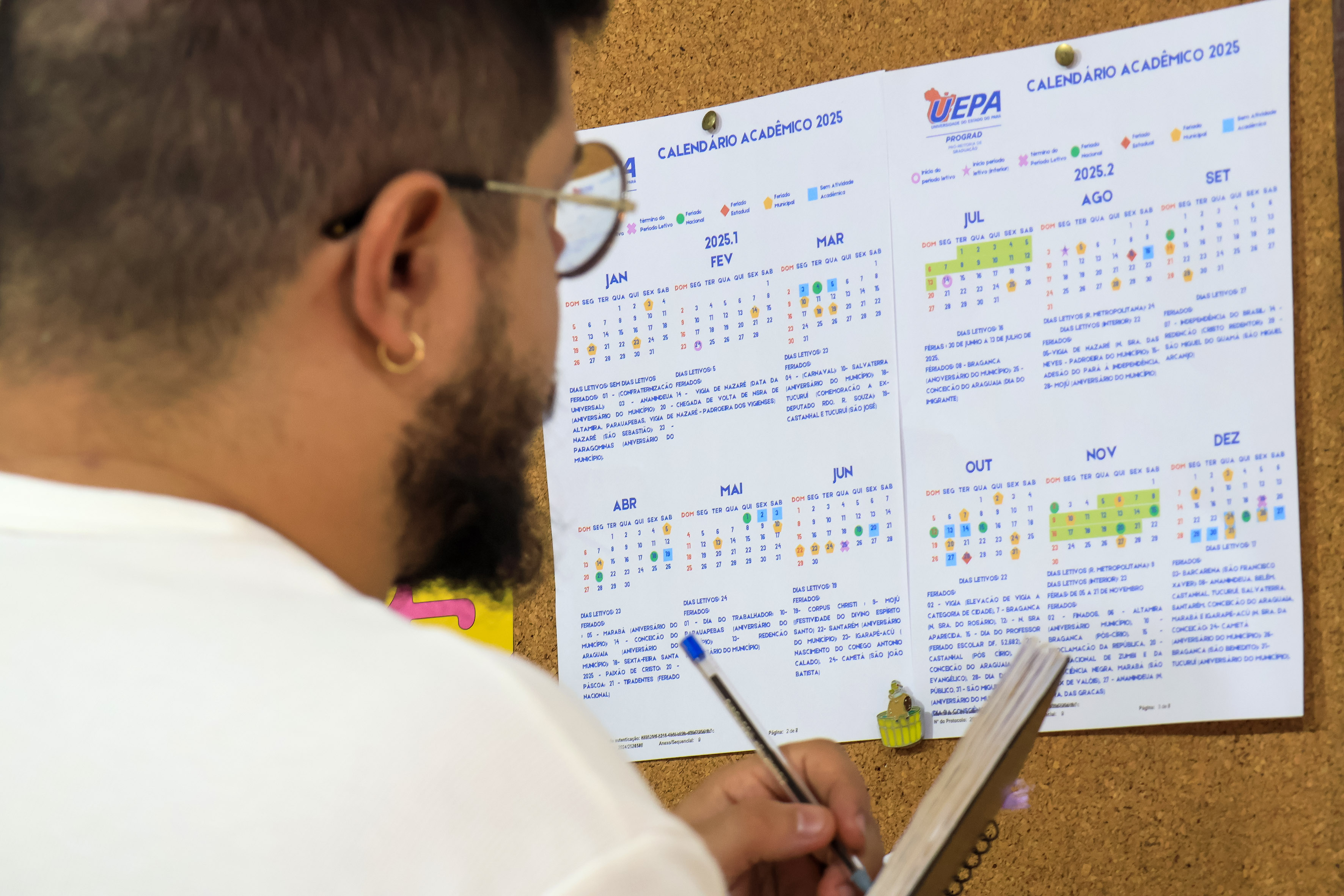 A imagem mostra uma pessoa, de costas e levemente de perfil, observando um calendário acadêmico fixado em um mural de cortiça. O calendário pertence à Universidade do Estado do Pará (Uepa) para o ano de 2025. A pessoa usa óculos e possui barba, além de um brinco de argola dourada. Ela está segurando uma caneta e um caderno, aparentemente anotando ou conferindo informações do calendário. O calendário apresenta meses divididos em colunas com legendas coloridas indicando eventos e períodos importantes.