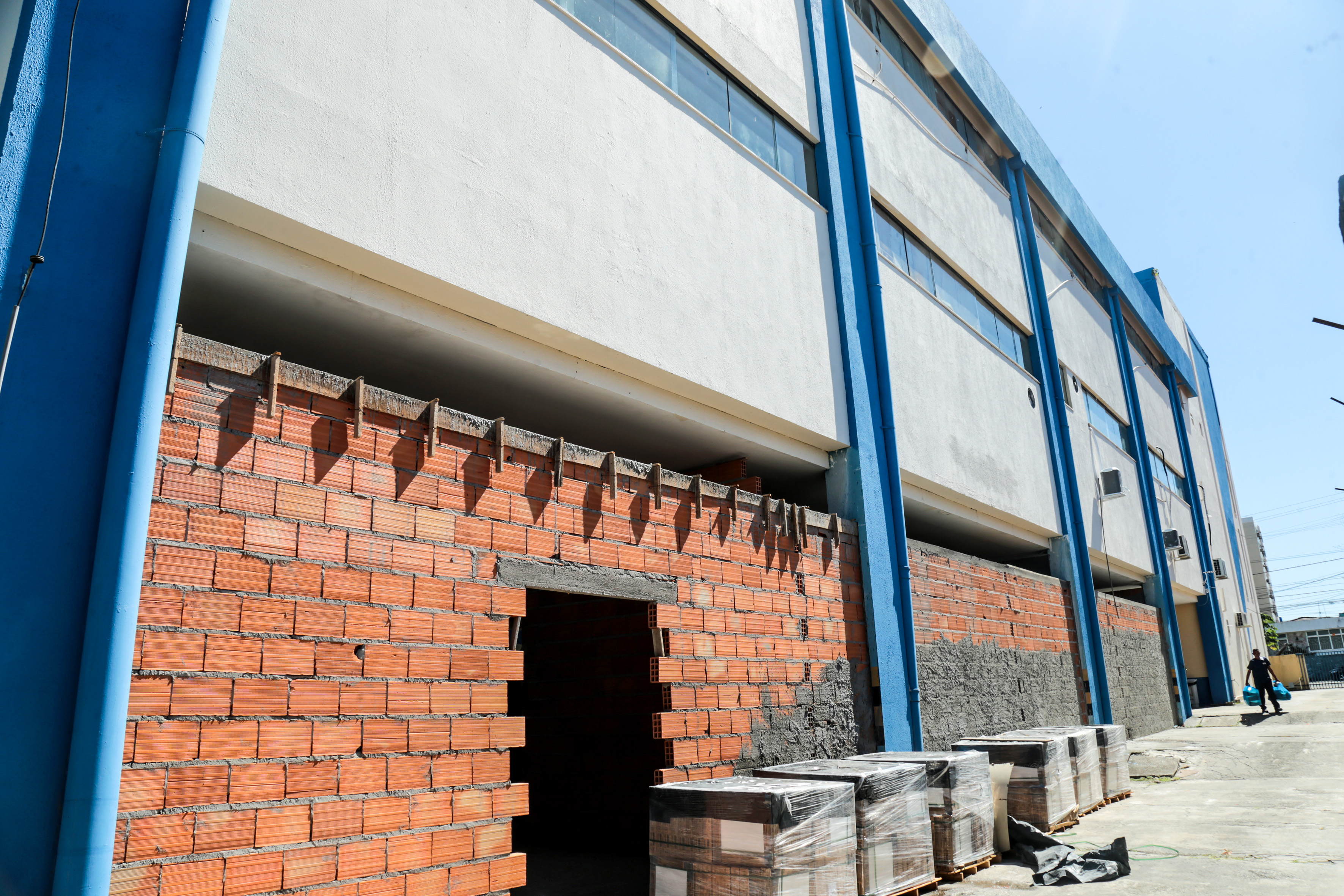 A imagem mostra a fachada exterior de um edifício com uma estrutura de porta metálica azul, com uma seção inacabada com tijolos vermelhos expostos e uma porta aberta sem porta, indicando trabalho de construção ou reparo em andamento. 